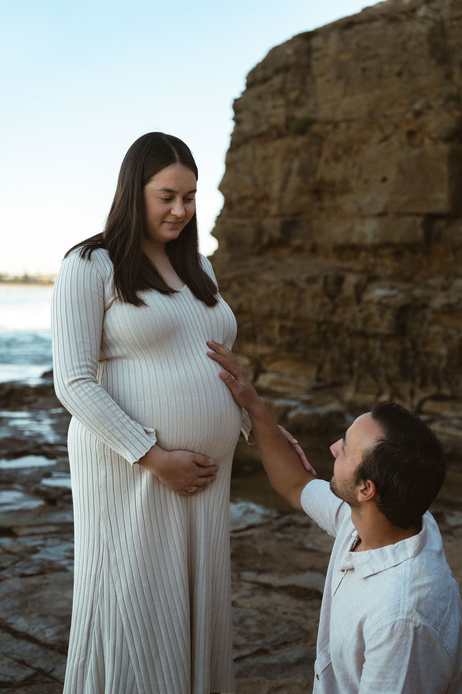 Pregnant woman in white dress with partner kneeling beside her on rocky beach at sunset.
