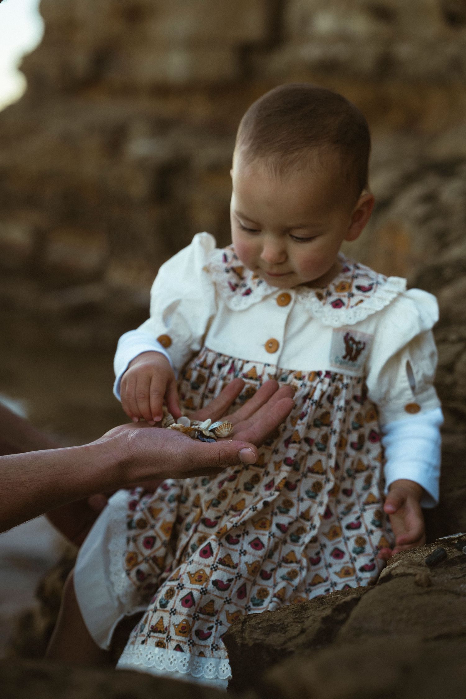 Baby in floral dress examines small objects in an adult's outstretched hand while sitting on rocks outdoors.