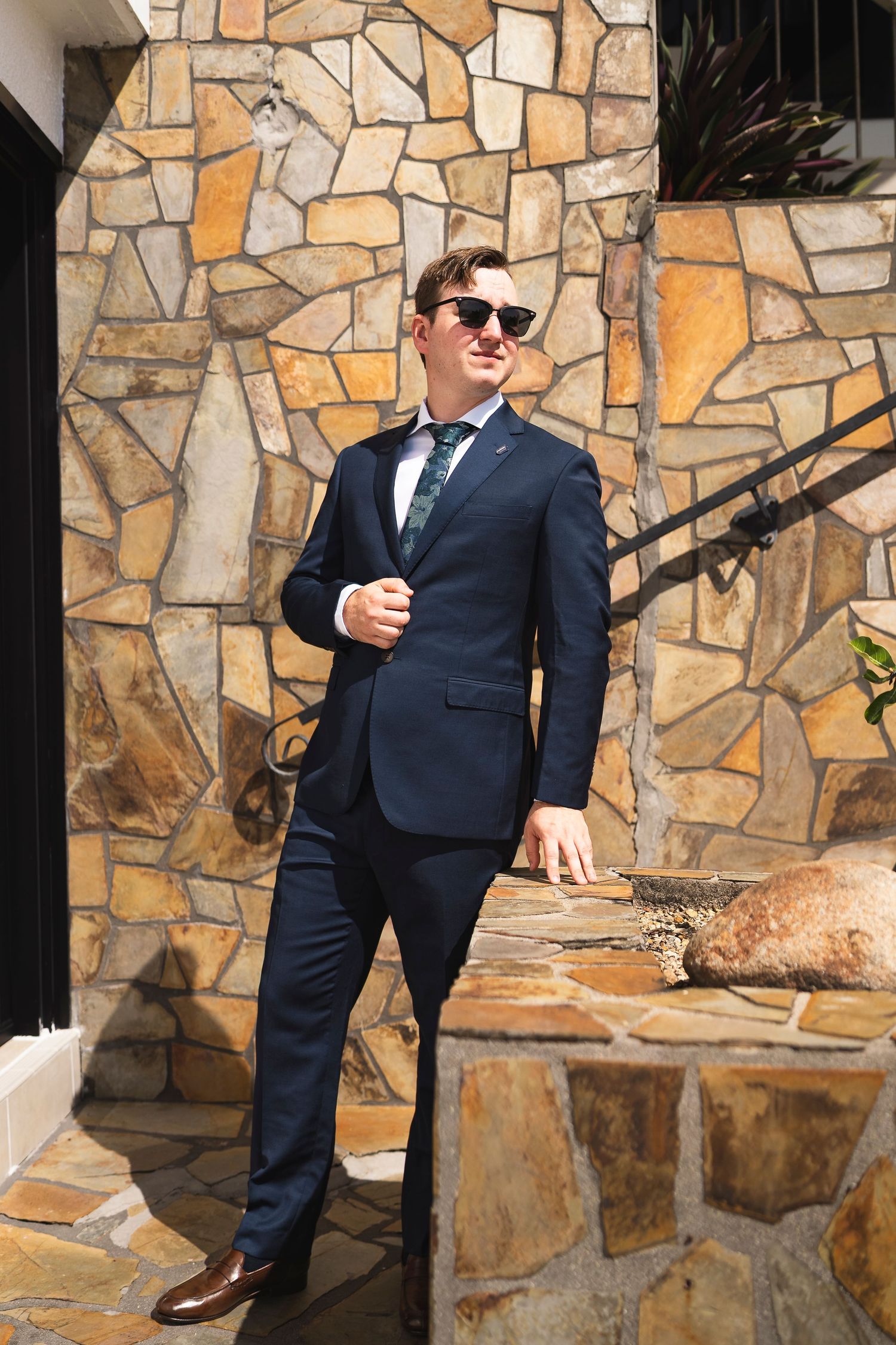 Man in navy blue suit and sunglasses posing confidently against a rustic stone wall outdoors.