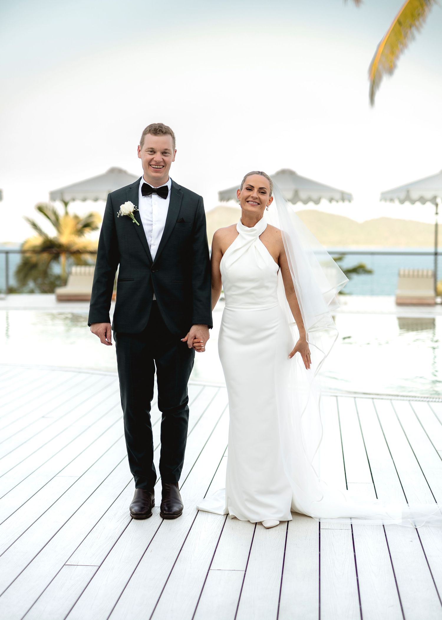 Bride in white halter gown and groom in black tuxedo hold hands smiling at outdoor waterfront wedding venue.