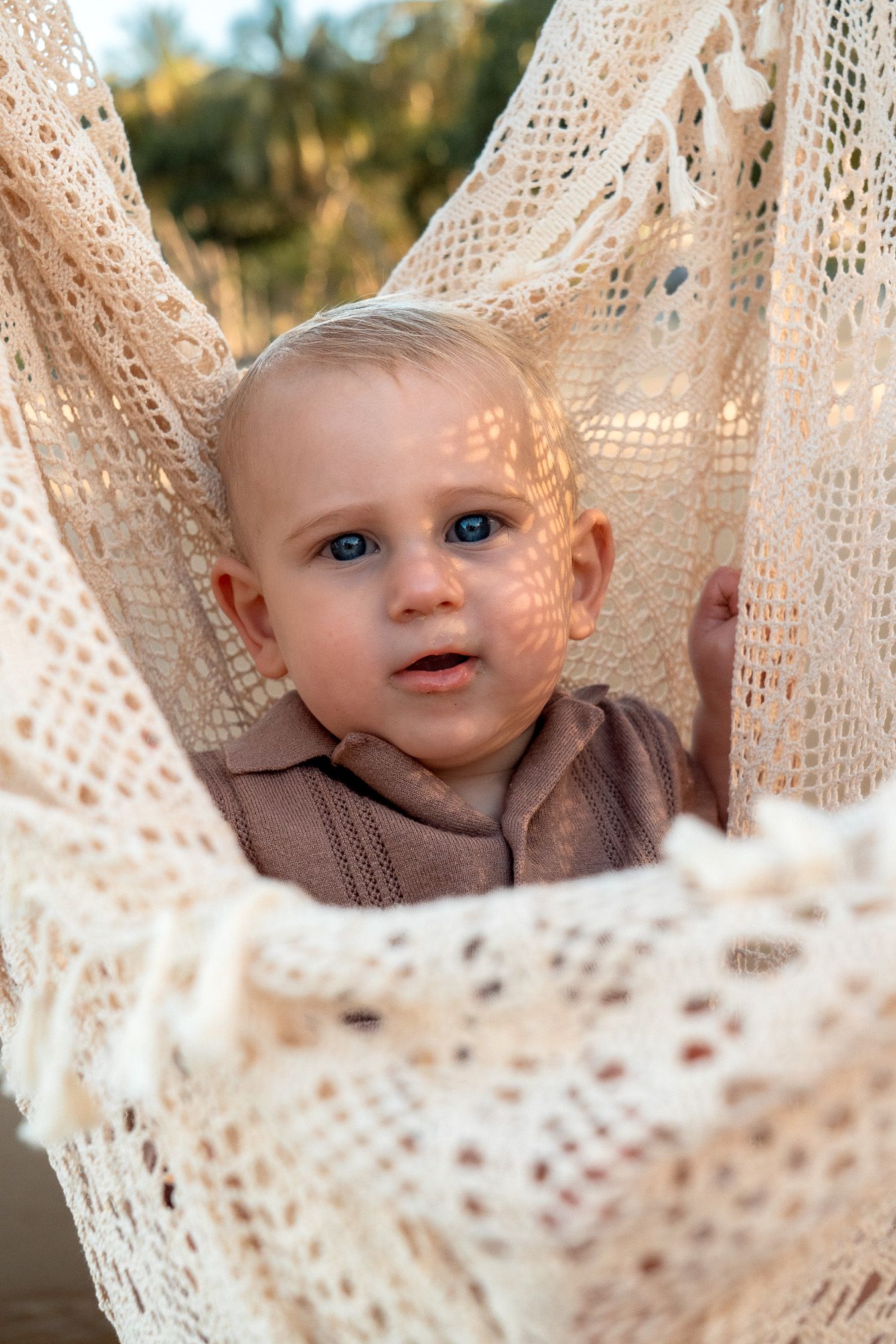 A blue-eyed baby sitting in a lace hammock with dappled sunlight outdoors.