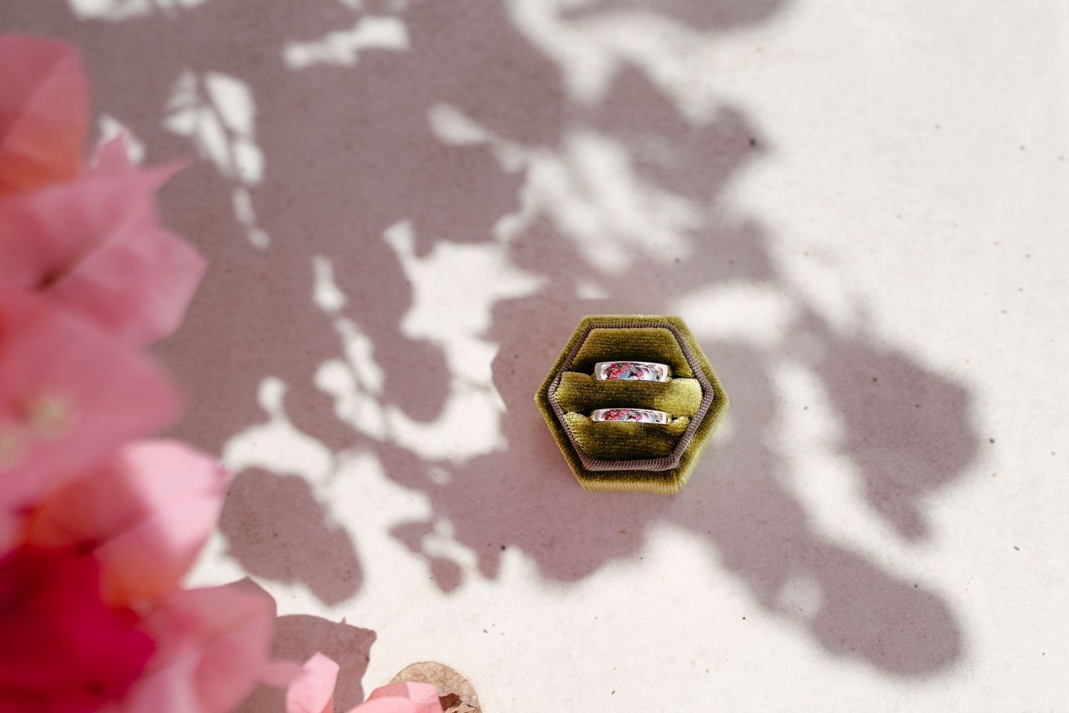 Gold hexagonal ring box holding two rings with pink gemstones, surrounded by pink flowers and soft shadows.