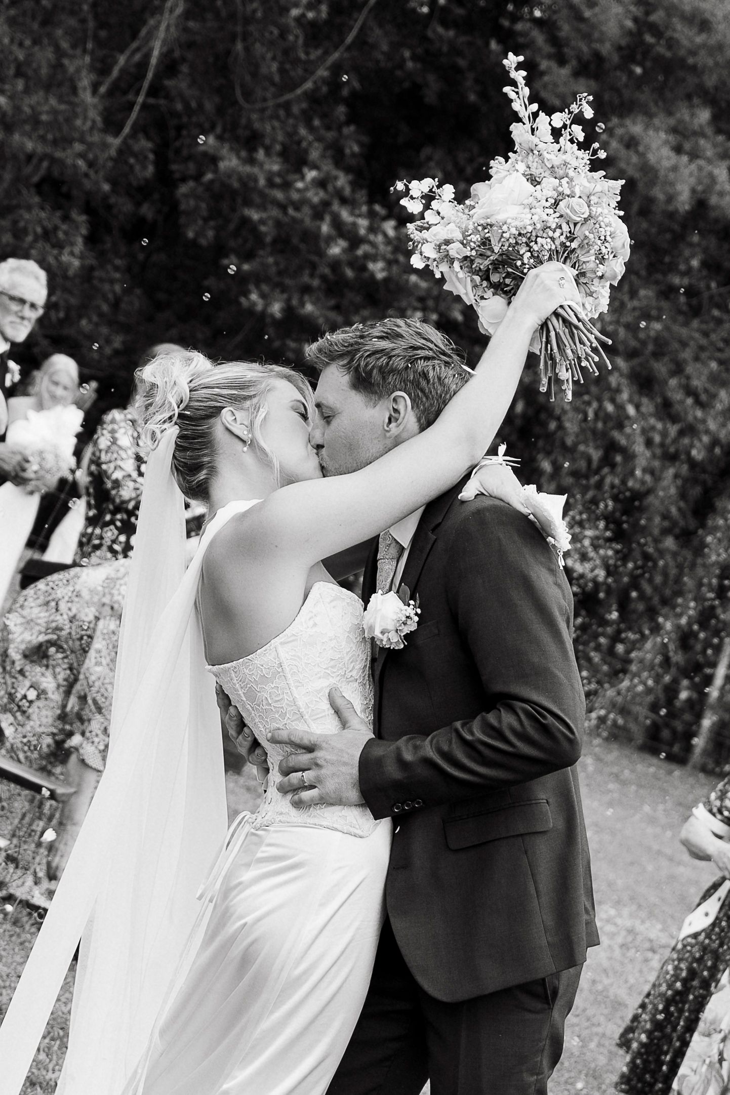 Black and white photo of a bride and groom sharing their first kiss, bride raising bouquet joyfully.