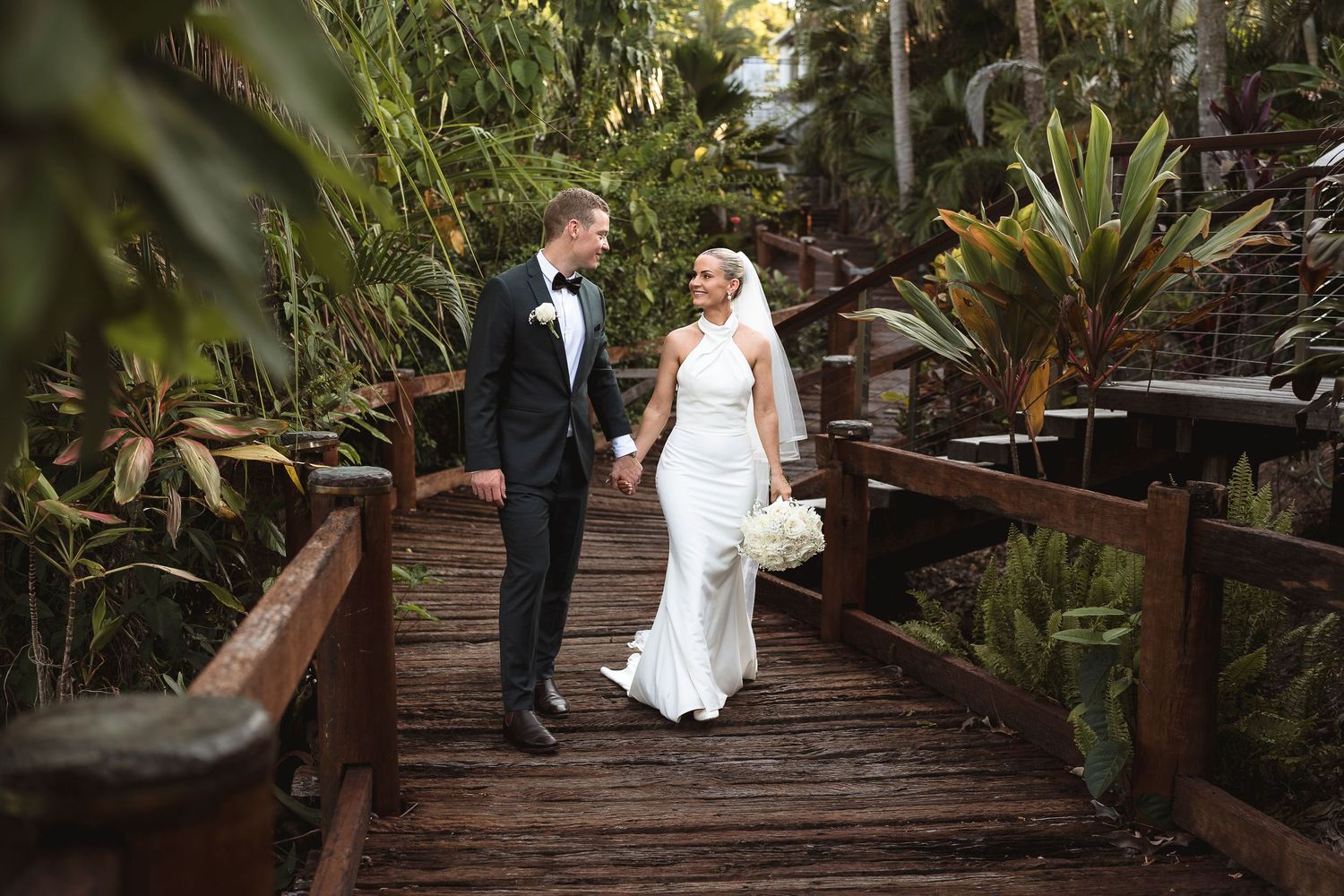 Bride and groom holding hands on a wooden bridge surrounded by lush tropical greenery on their wedding day.