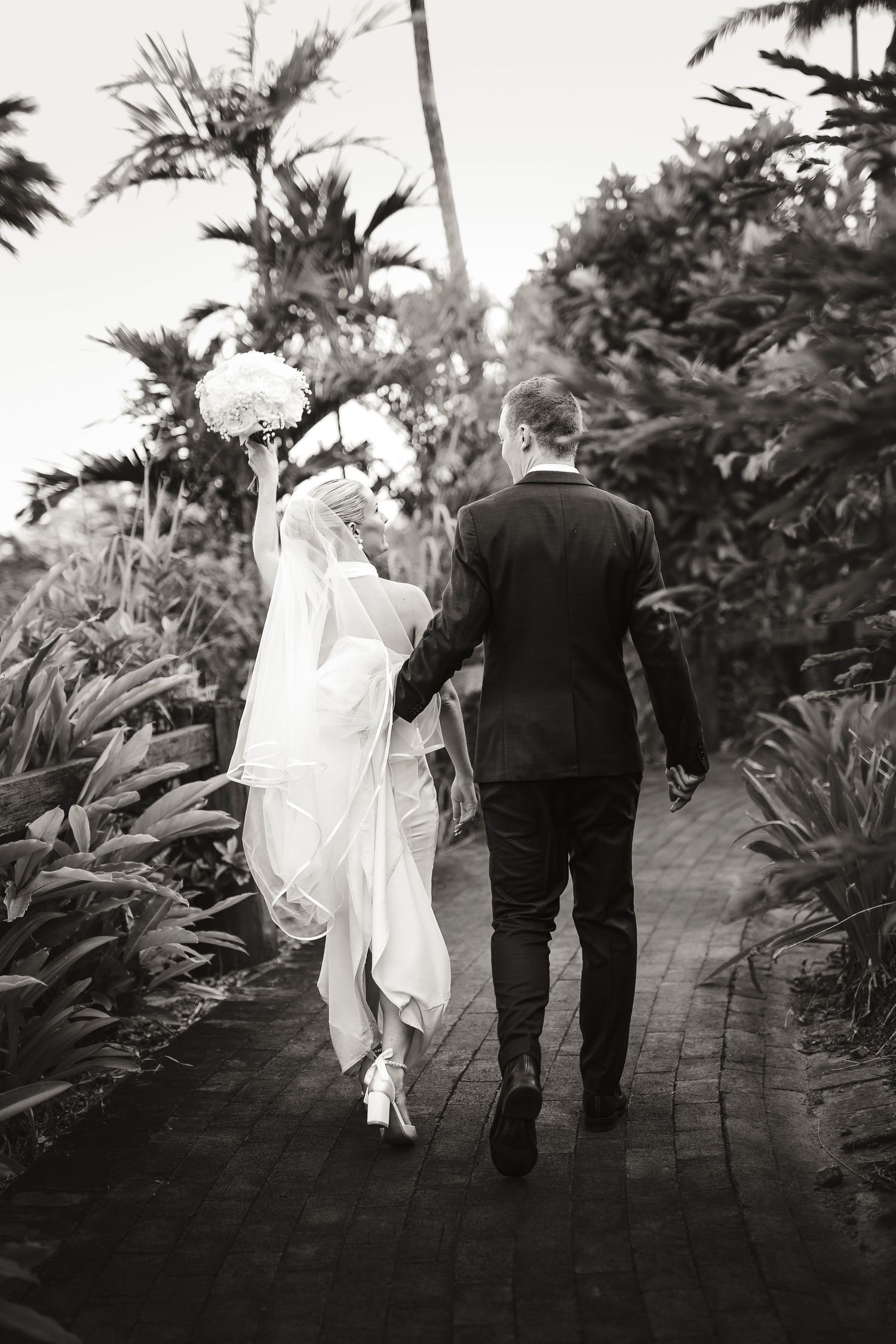 Black and white photo of bride and groom walking hand in hand down a tropical garden path, bride raising bouquet.
