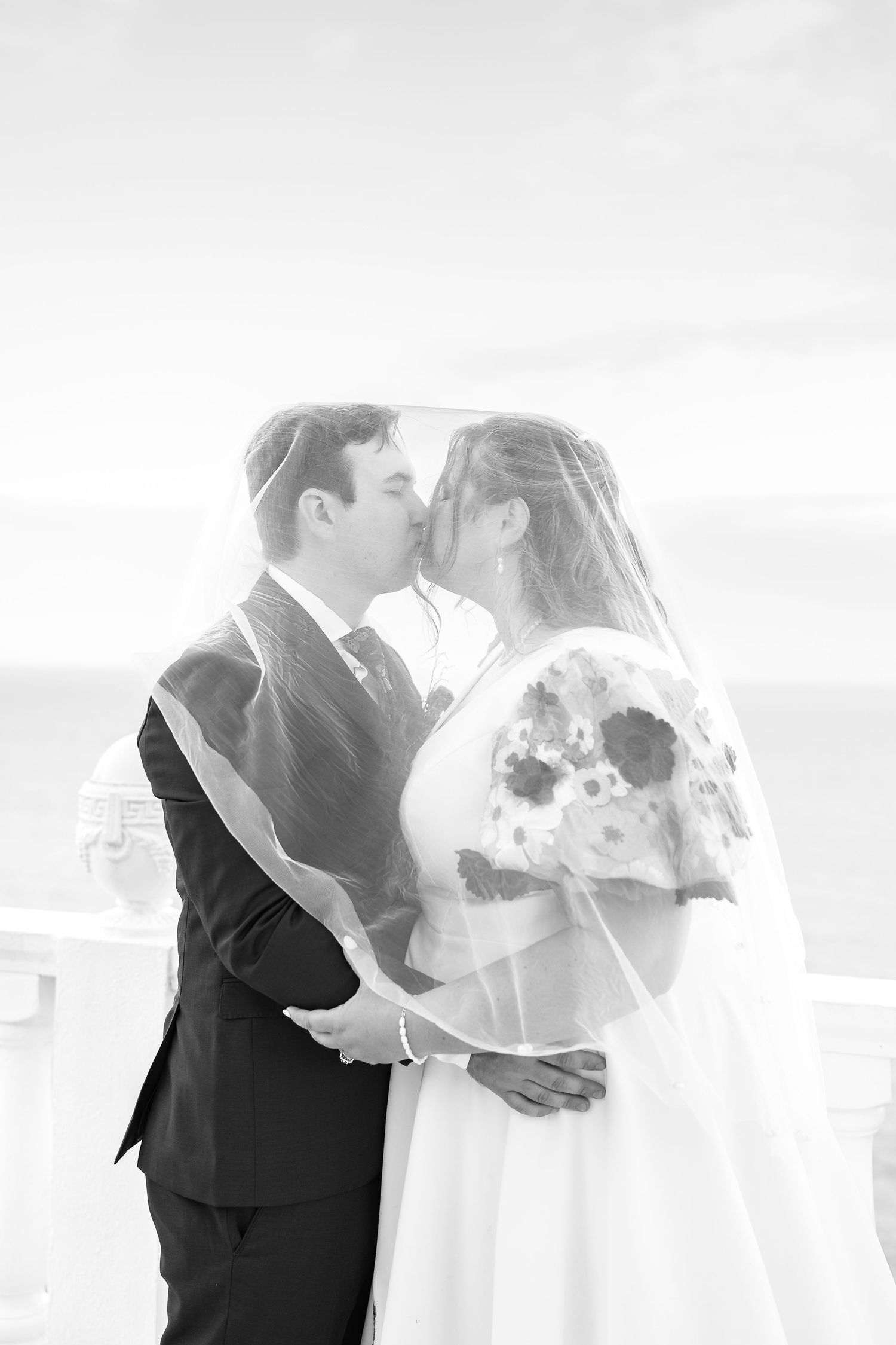 Black and white photo of bride and groom kissing under veil outdoors near ocean on wedding day.