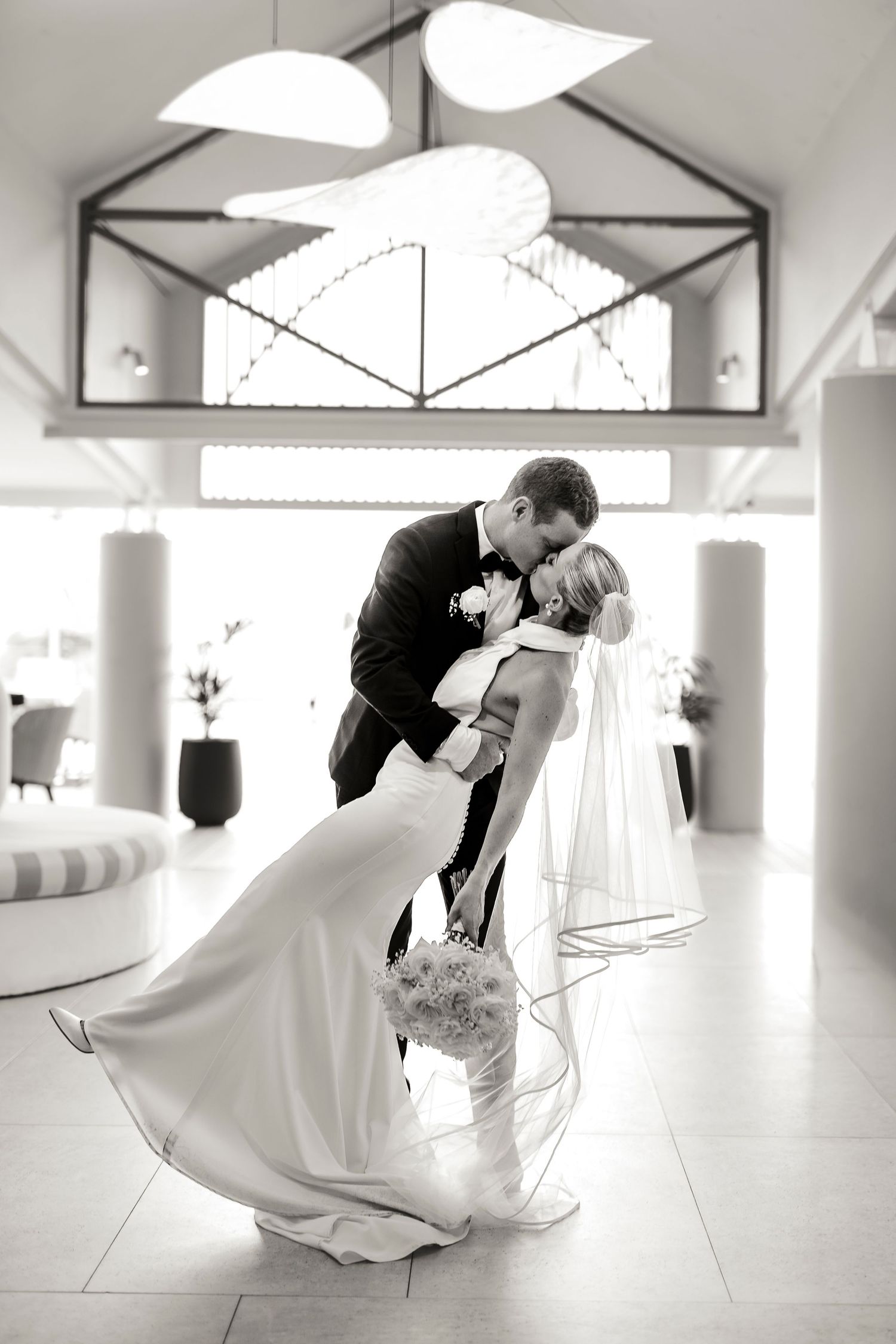 Black and white wedding photo of groom dipping bride in elegant venue, her veil flowing as they share a romantic kiss.