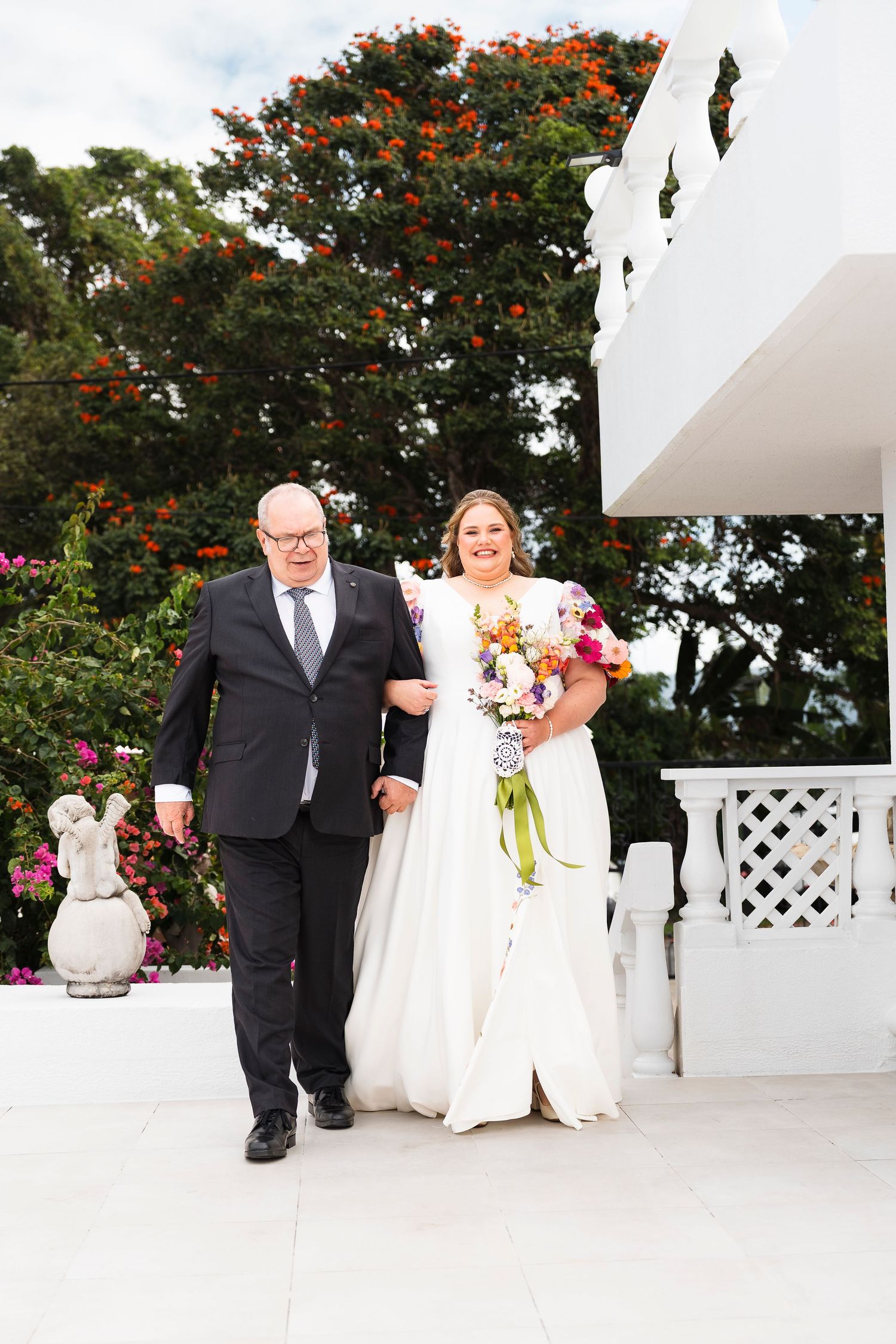 Bride in white gown holding colorful bouquet walks with older man in black suit at outdoor wedding venue.