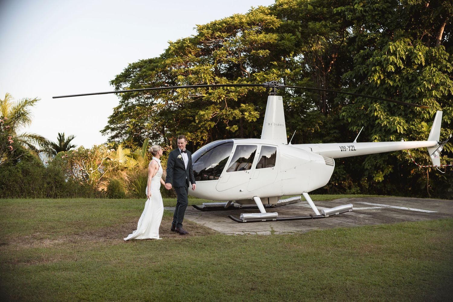 Bride and groom posing beside a white helicopter on a lush green lawn at their outdoor wedding.