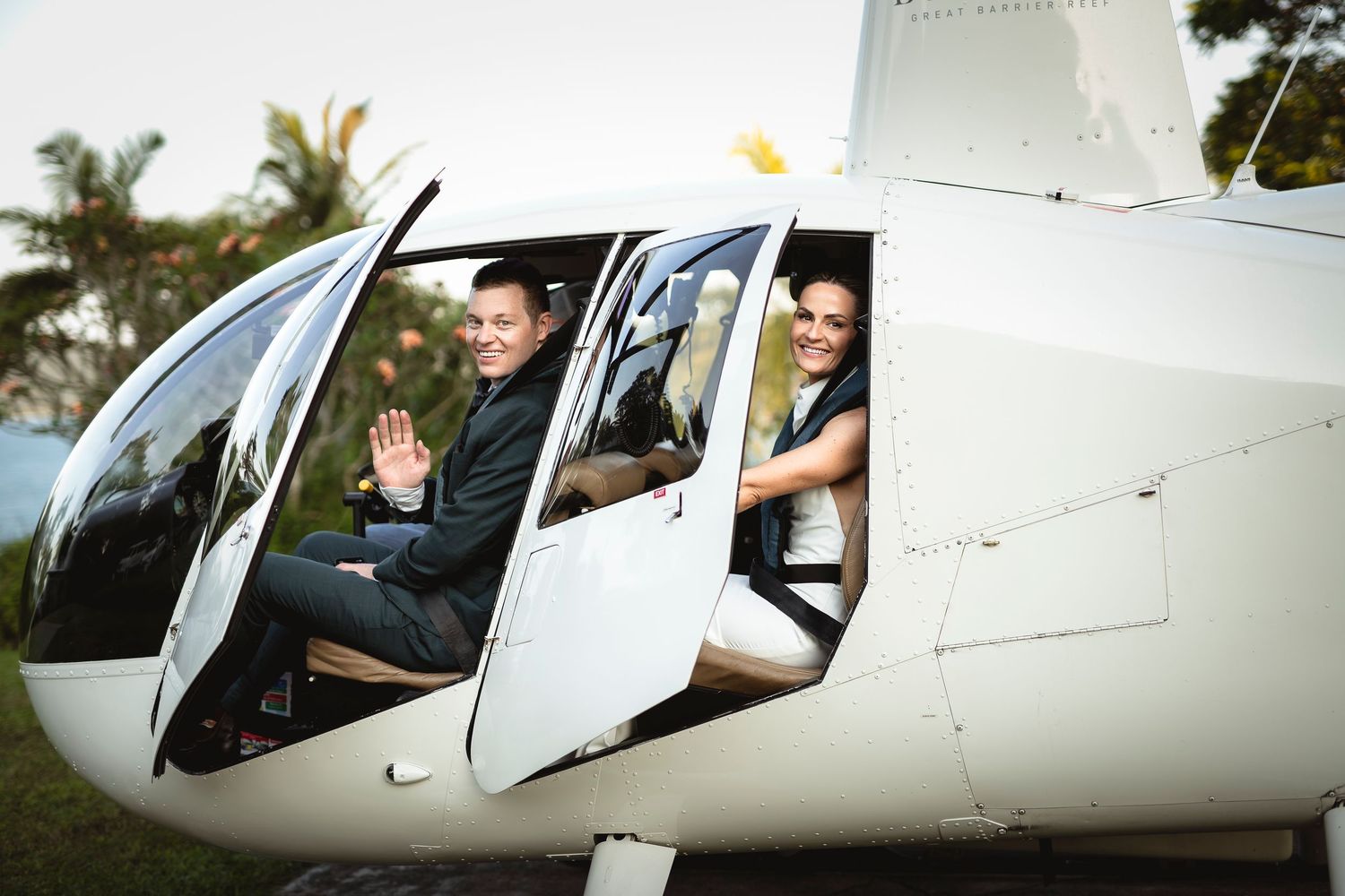 Smiling couple in formal attire waving from a white helicopter cockpit with tropical trees in background.
