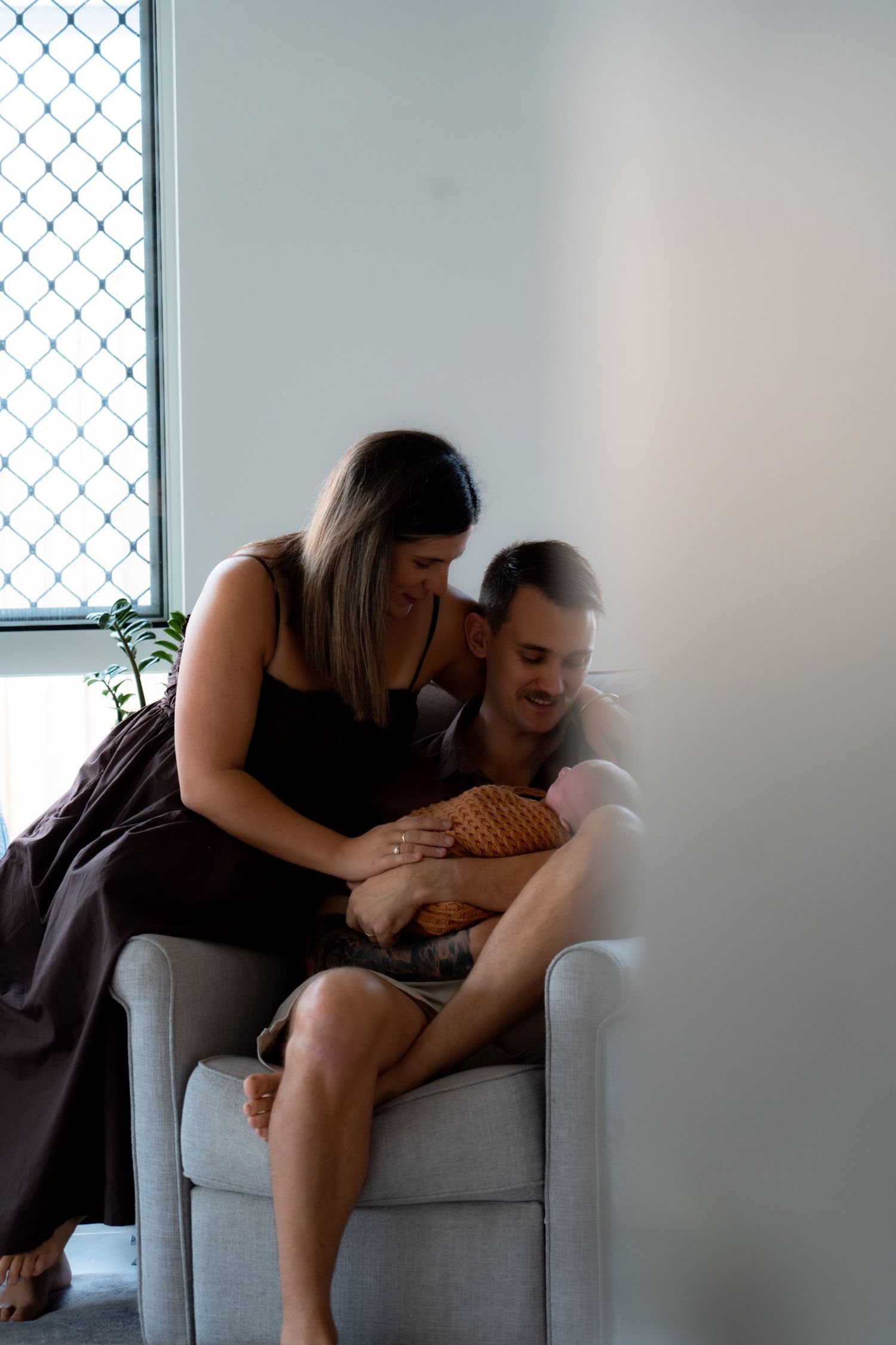 New parents sitting in armchair by window lovingly gazing at their newborn baby together.