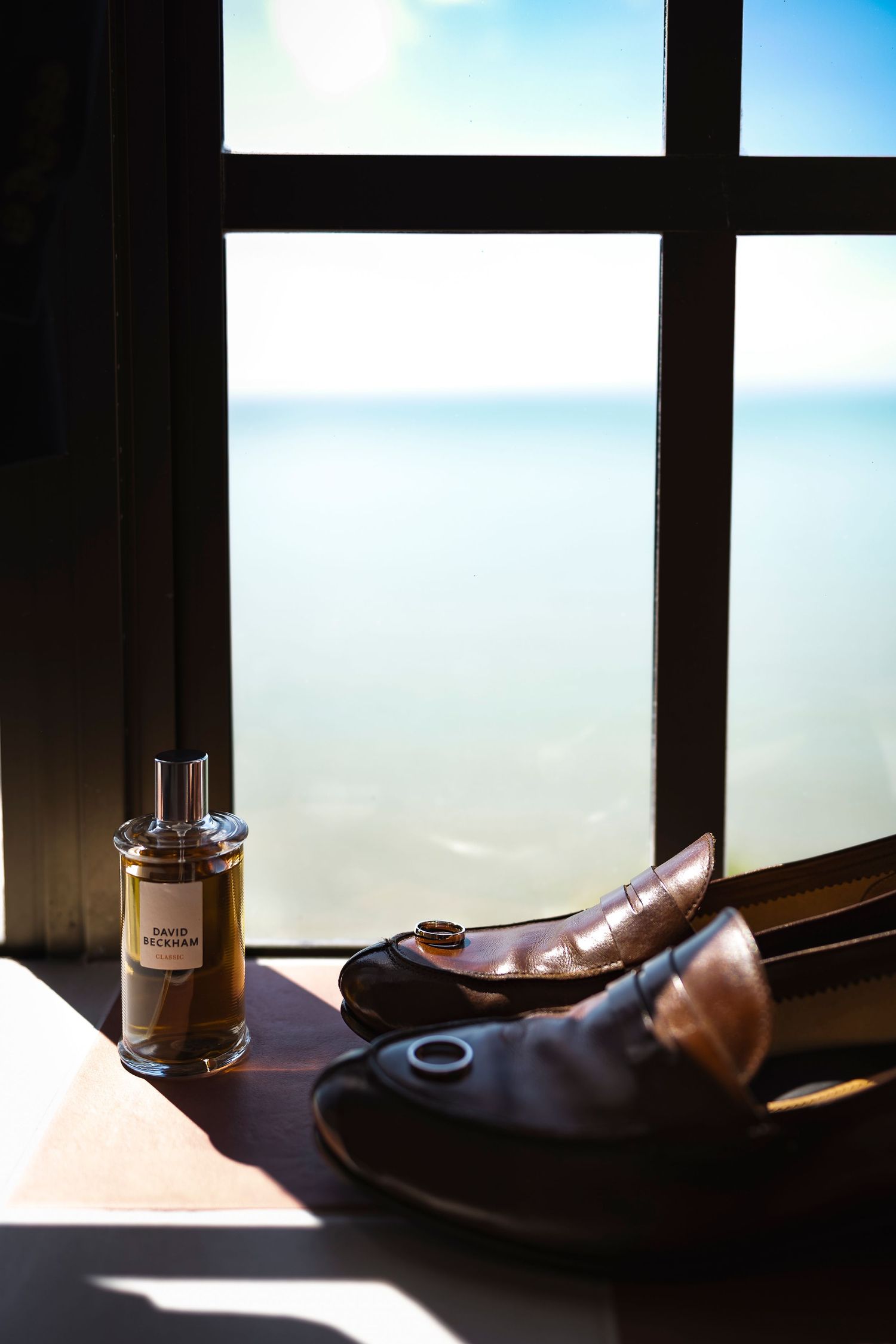 Brown leather loafers and a cologne bottle on a wooden surface by a window overlooking a serene ocean view.