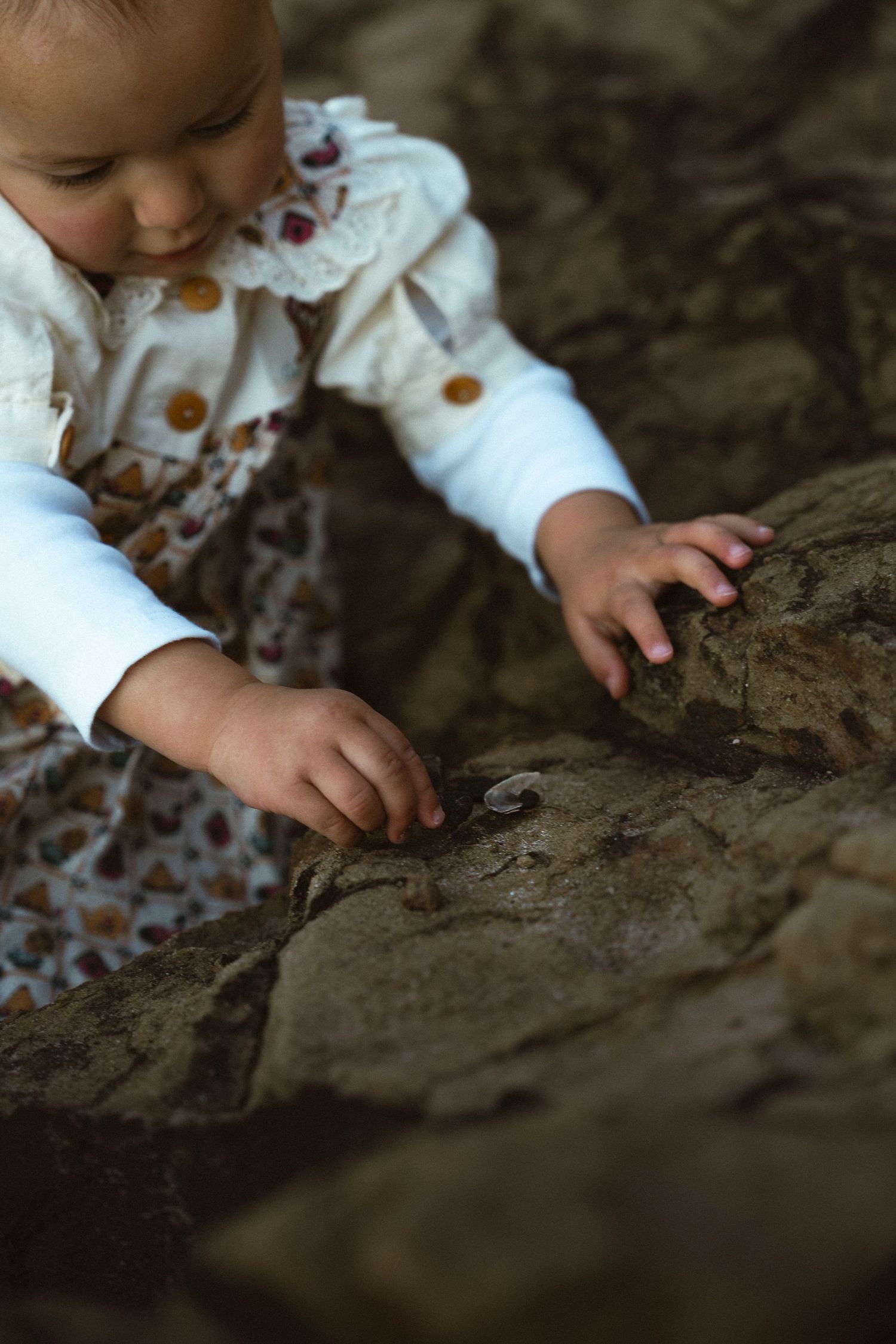 A curious baby in a floral dress explores rocky terrain, reaching out tiny hands to touch the textured stone surface.