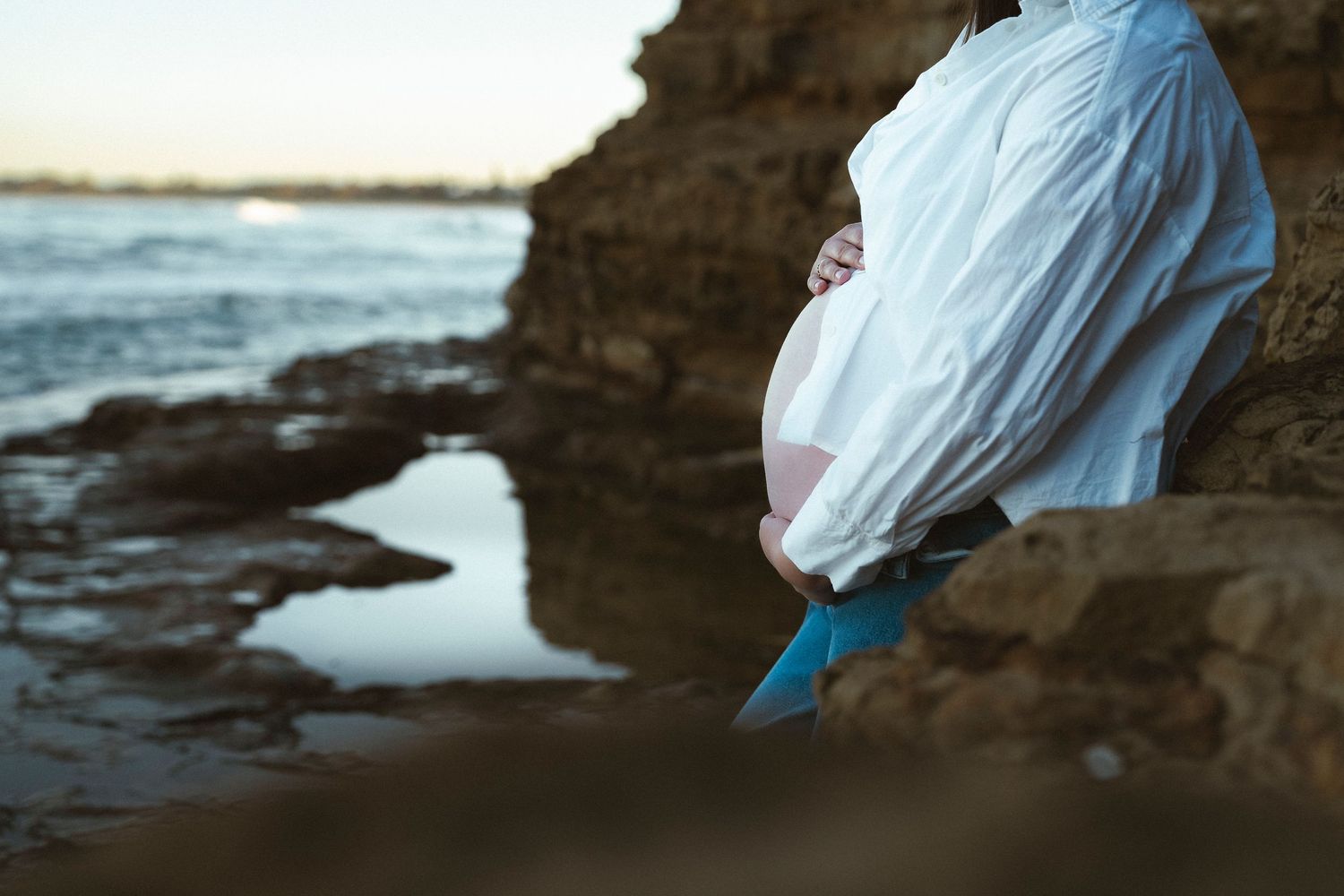 Pregnant woman draped in white fabric poses against rocky coastal cliffs with ocean waves in the background.