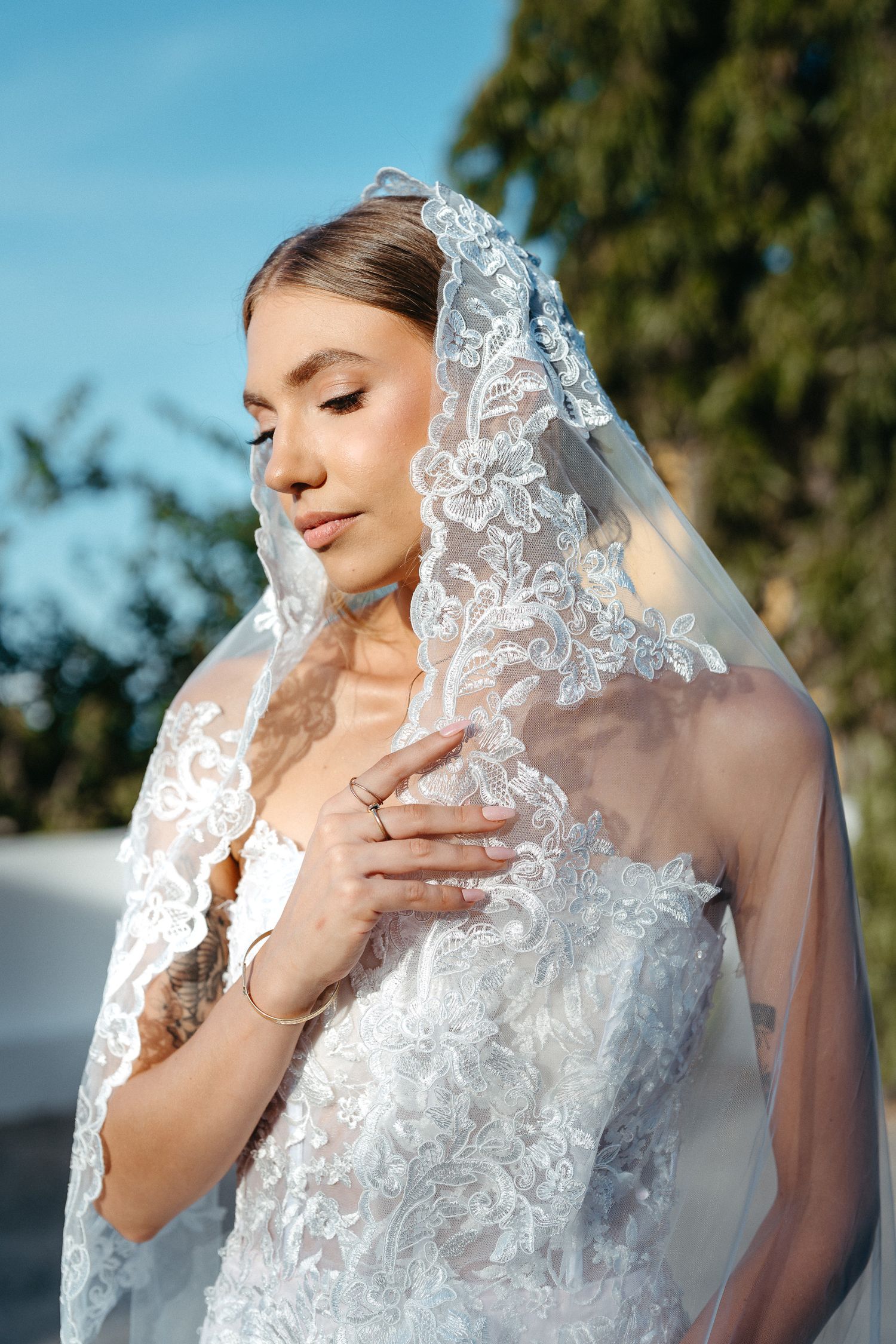 Bride in lace wedding dress and veil poses outdoors with eyes closed, hand on chest, trees in background.