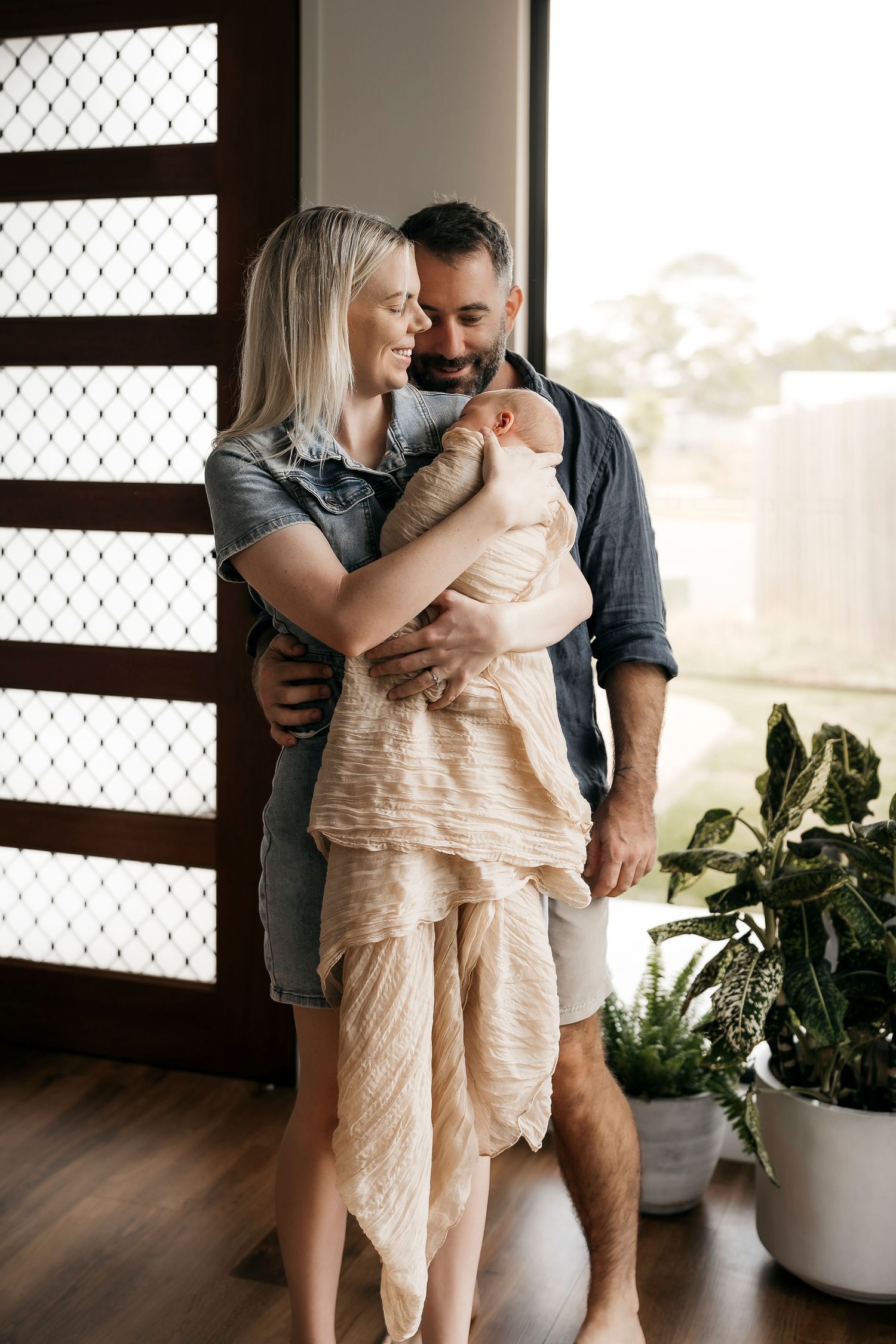 Happy couple standing by window smiling at their newborn baby wrapped in peach muslin.