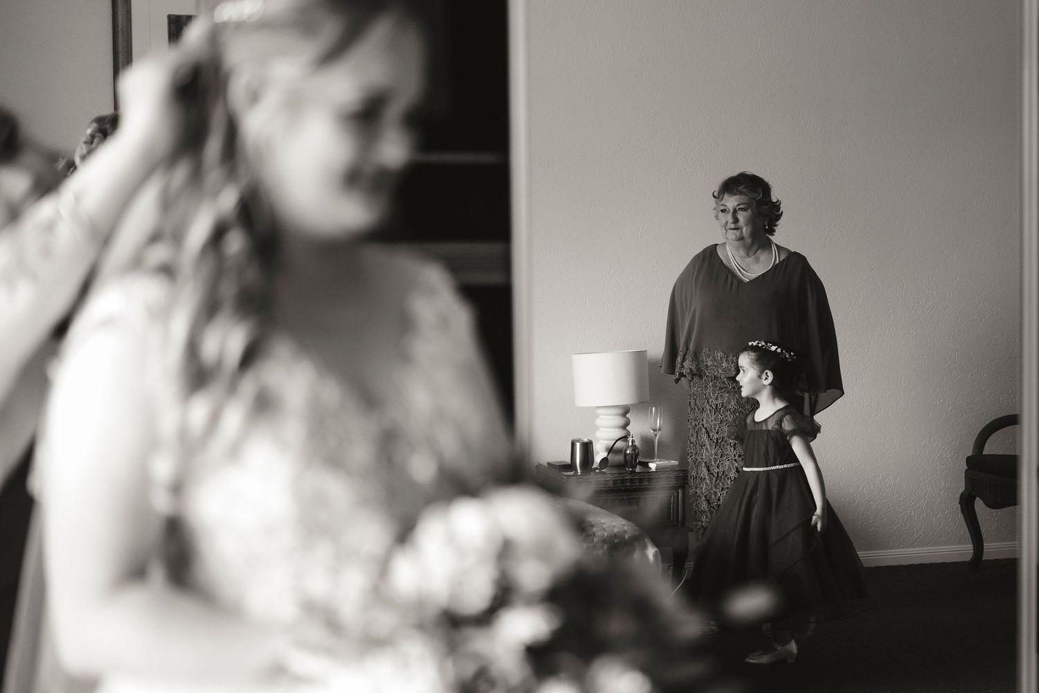 Black and white wedding photo of bride in foreground with older woman and flower girl watching in background.