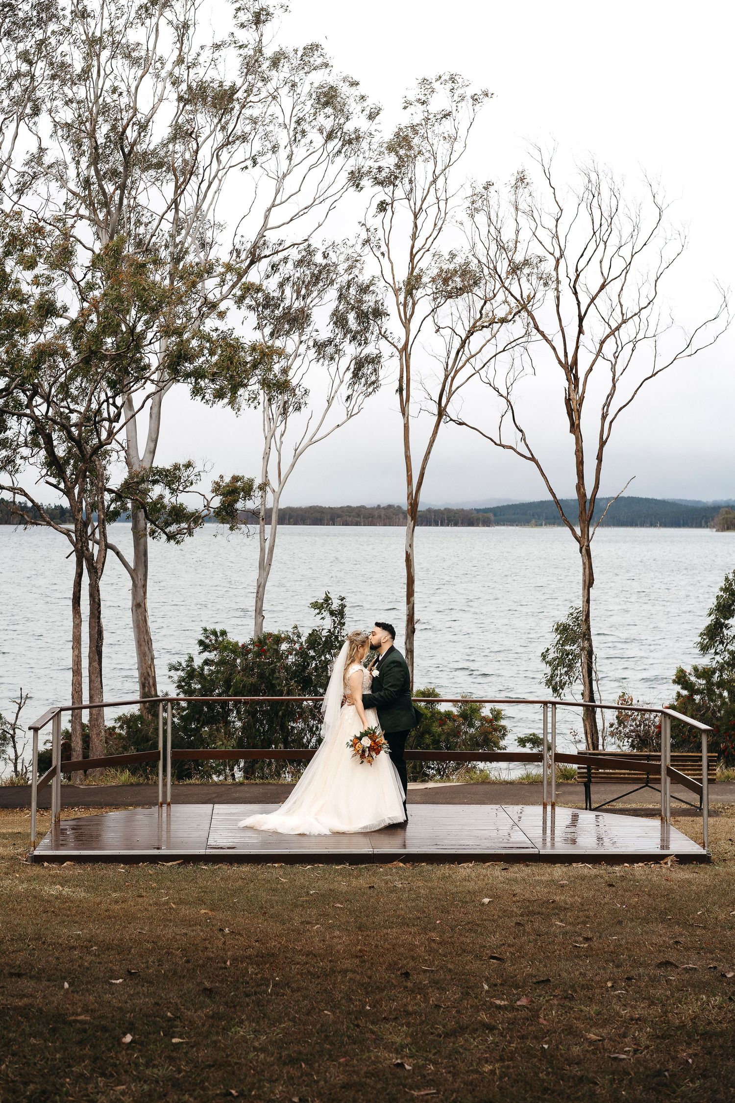 Bride and groom kissing on a lakeside platform surrounded by bare trees on a cloudy wedding day.