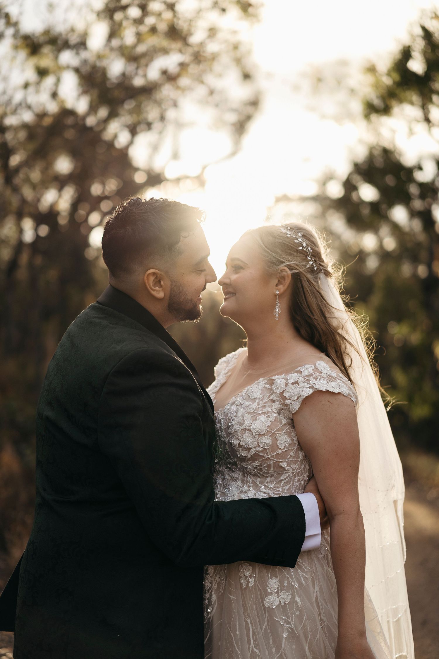 Bride and groom share a romantic moment outdoors, bathed in golden sunlight, with the bride in a lace gown and veil.
