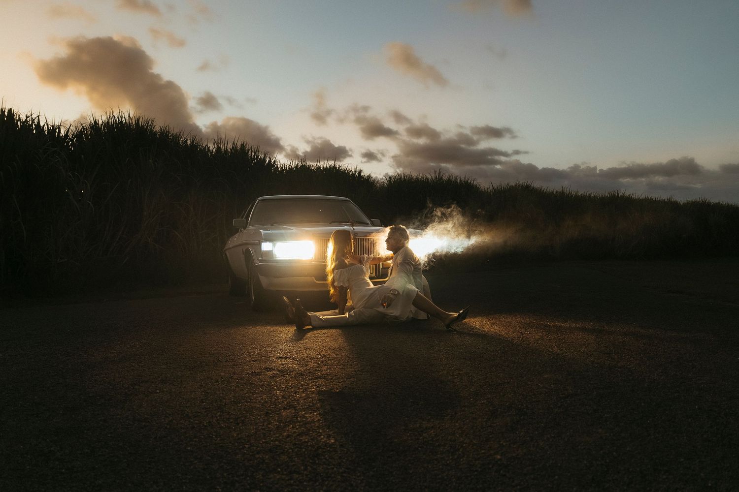 Couple sitting in front of vintage truck headlights at sunset on a rural road, romantic wedding photo.