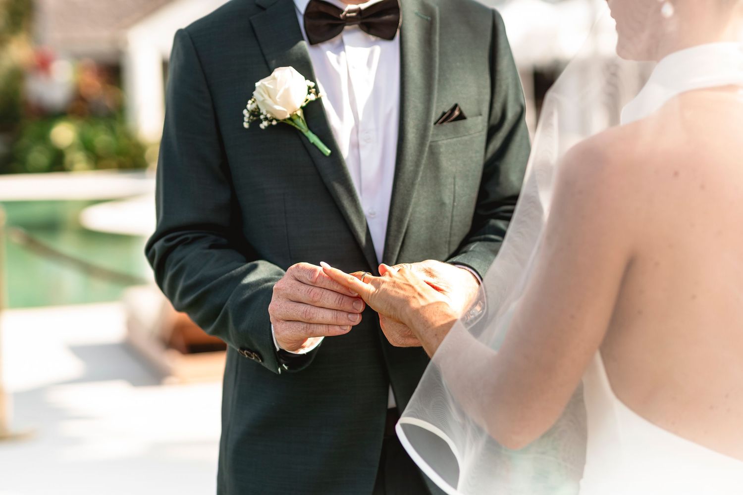 Groom in dark suit placing wedding ring on bride's finger during outdoor ceremony.