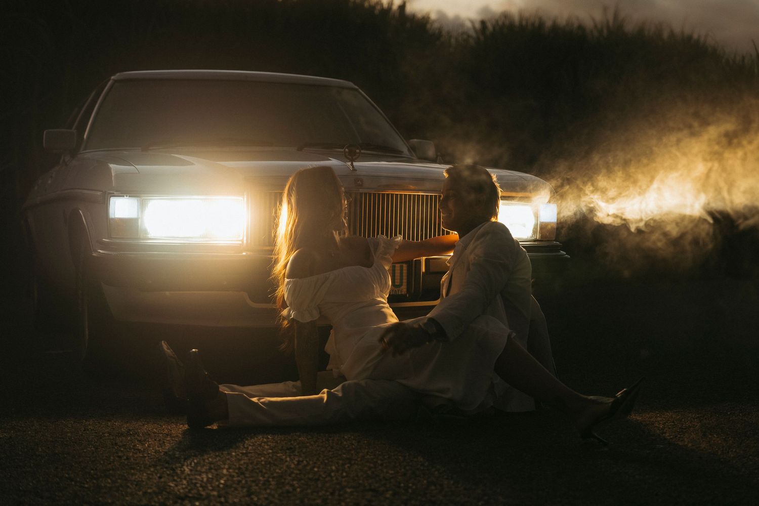 Two people sitting on ground in front of glowing car headlights at night with dramatic smoke and moody lighting.
