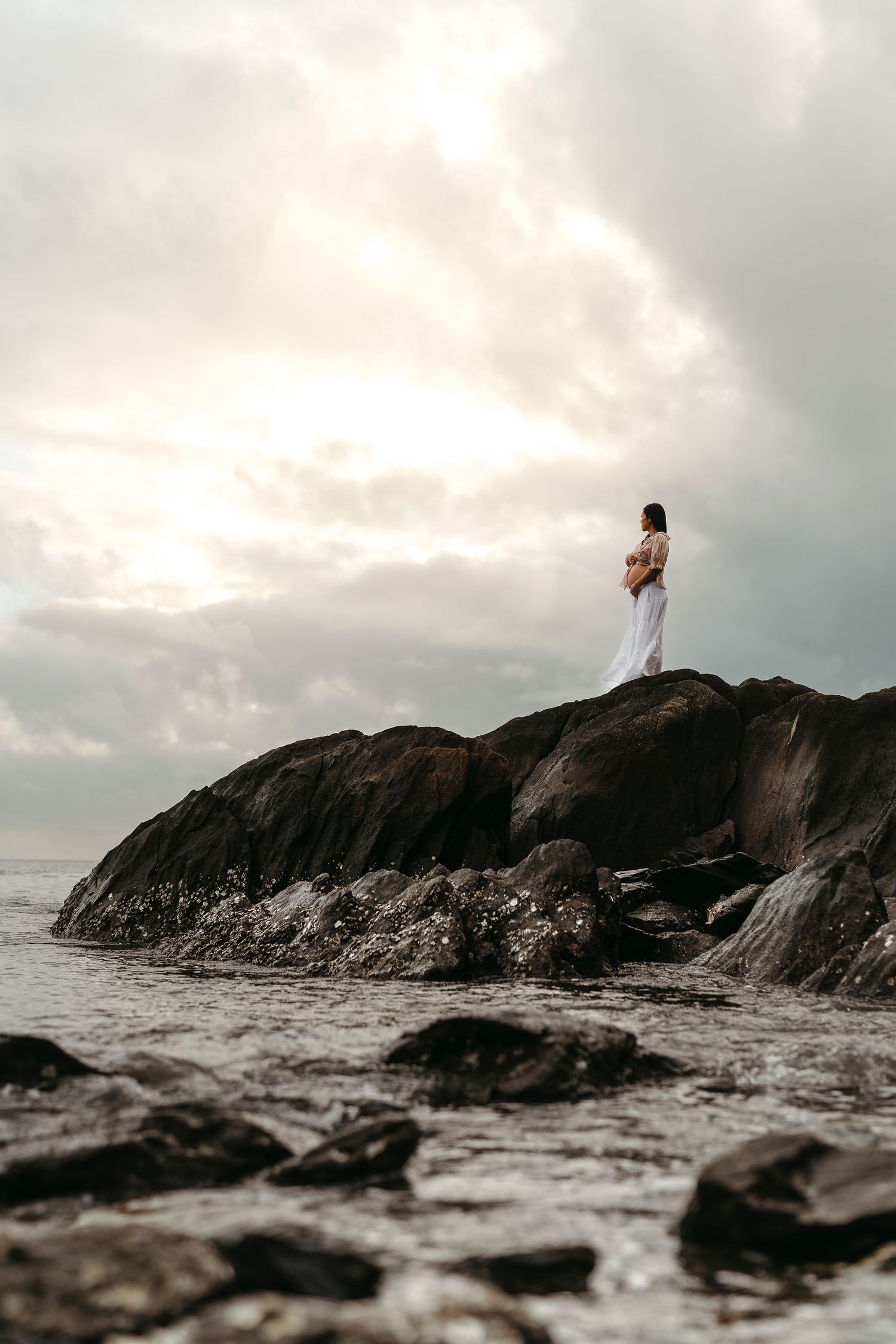 A woman in a white dress stands on rocky coastal cliffs overlooking a dramatic cloudy seascape at dusk.