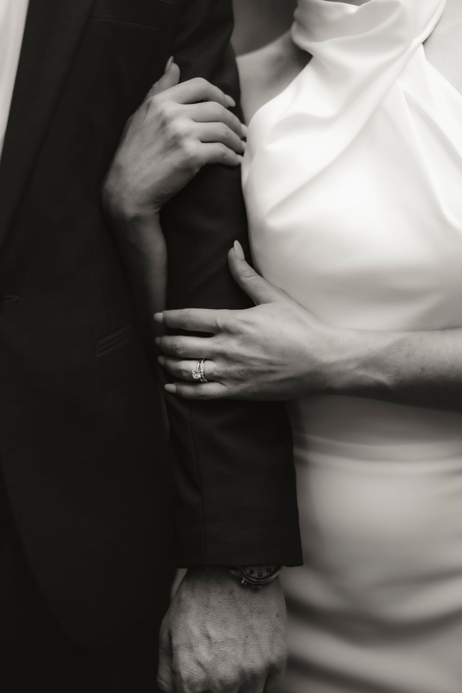 Black and white photo of a bride and groom embracing, showcasing her diamond engagement ring and wedding dress.