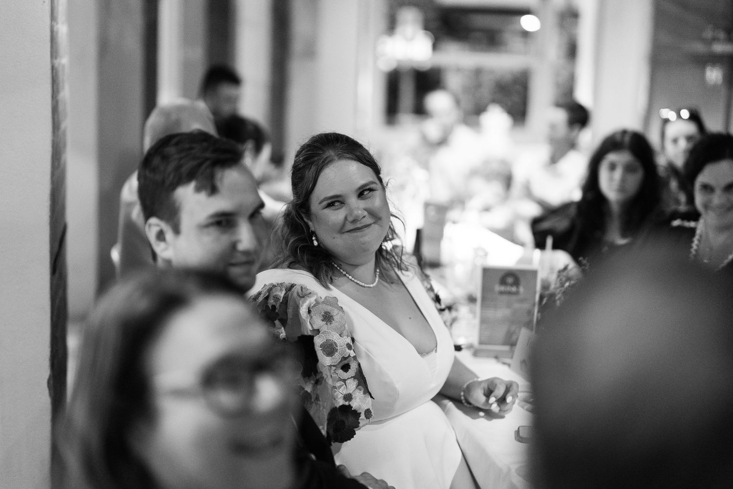 Black and white wedding reception photo of smiling bride in white dress seated among guests at elegant venue.