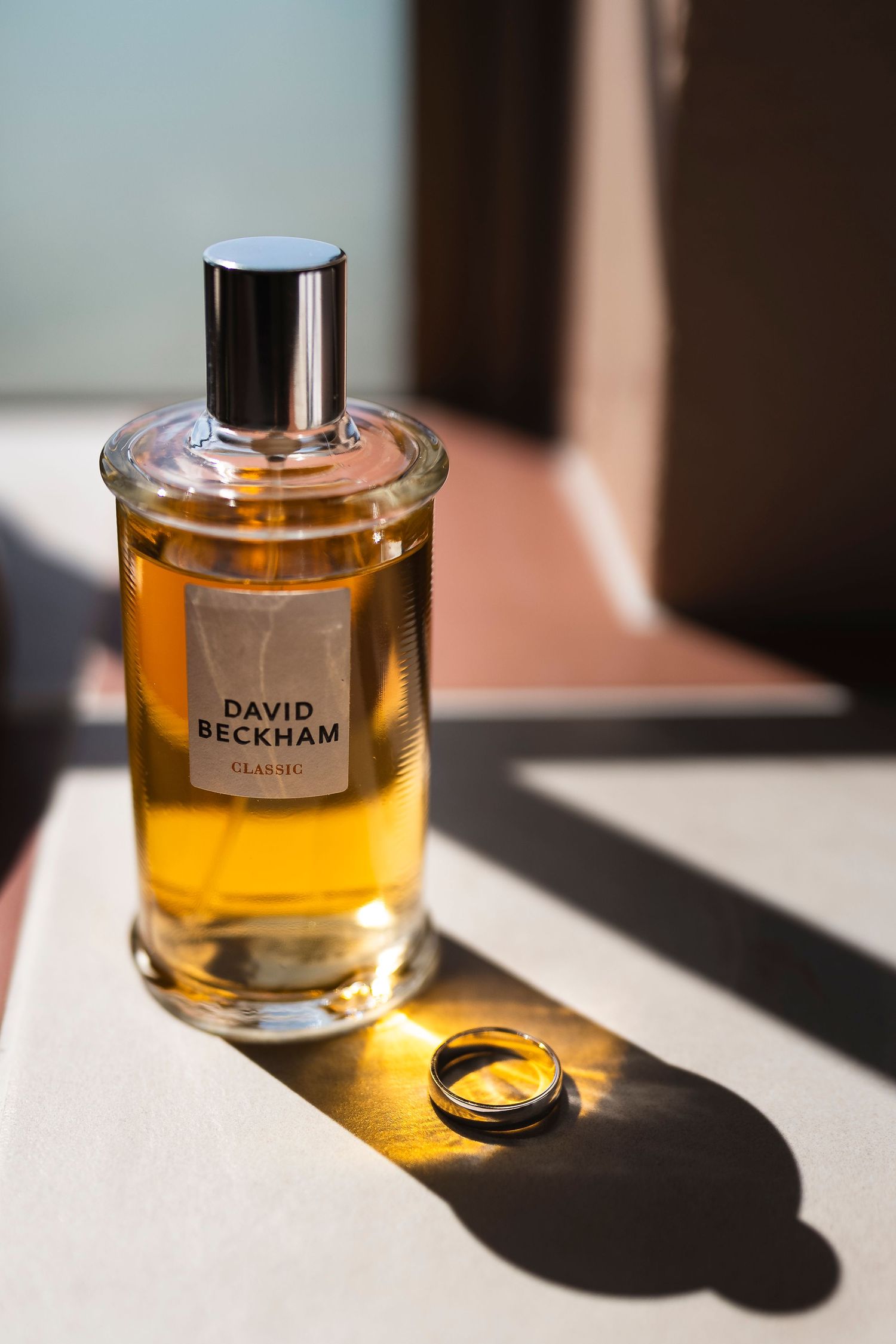 David Beckham Classic cologne bottle with gold ring beside it on a white surface in warm sunlight.