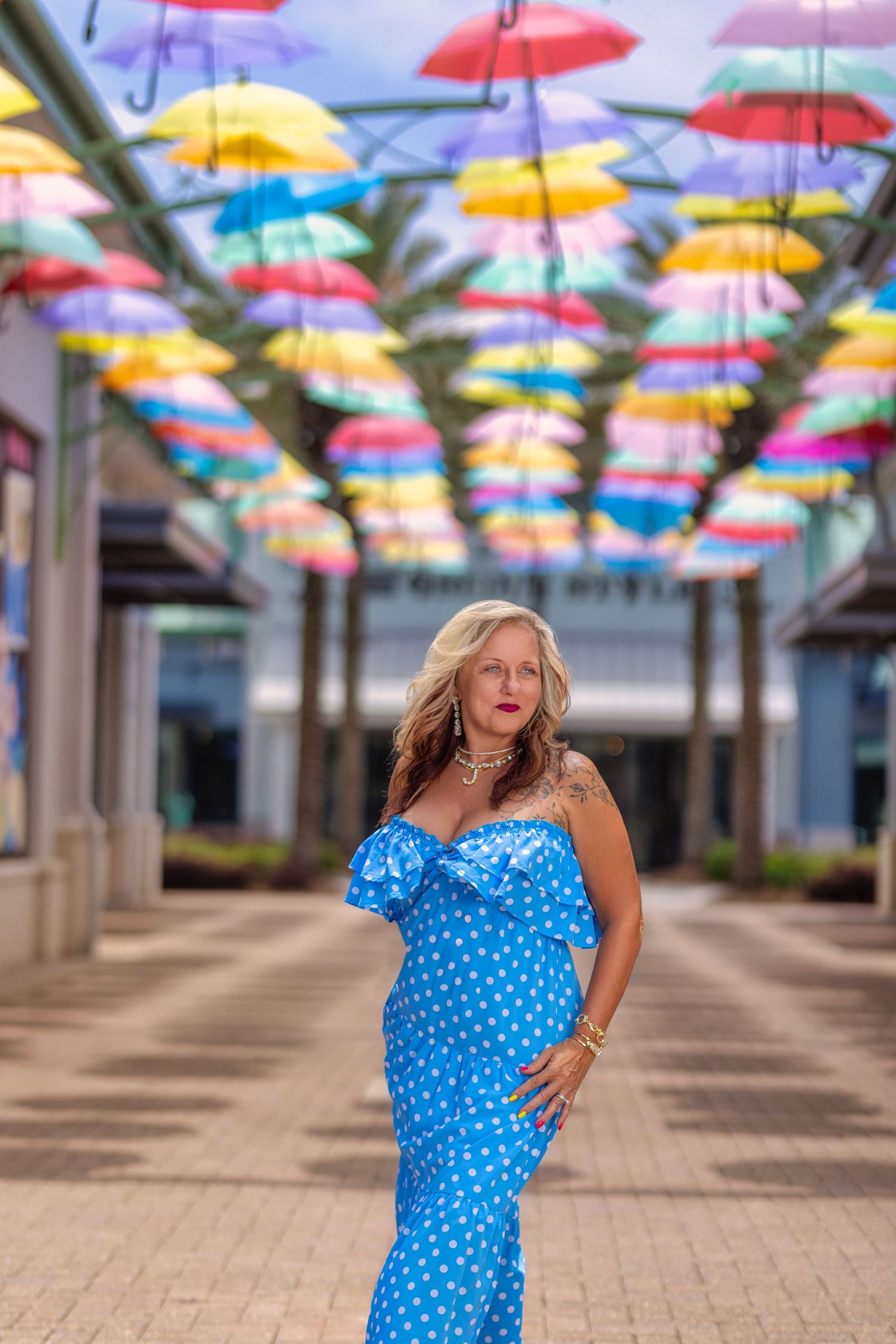 Someone poses in a polka dot blue dress beneath a colorful canopy of hanging umbrellas in an outdoor plaza area.