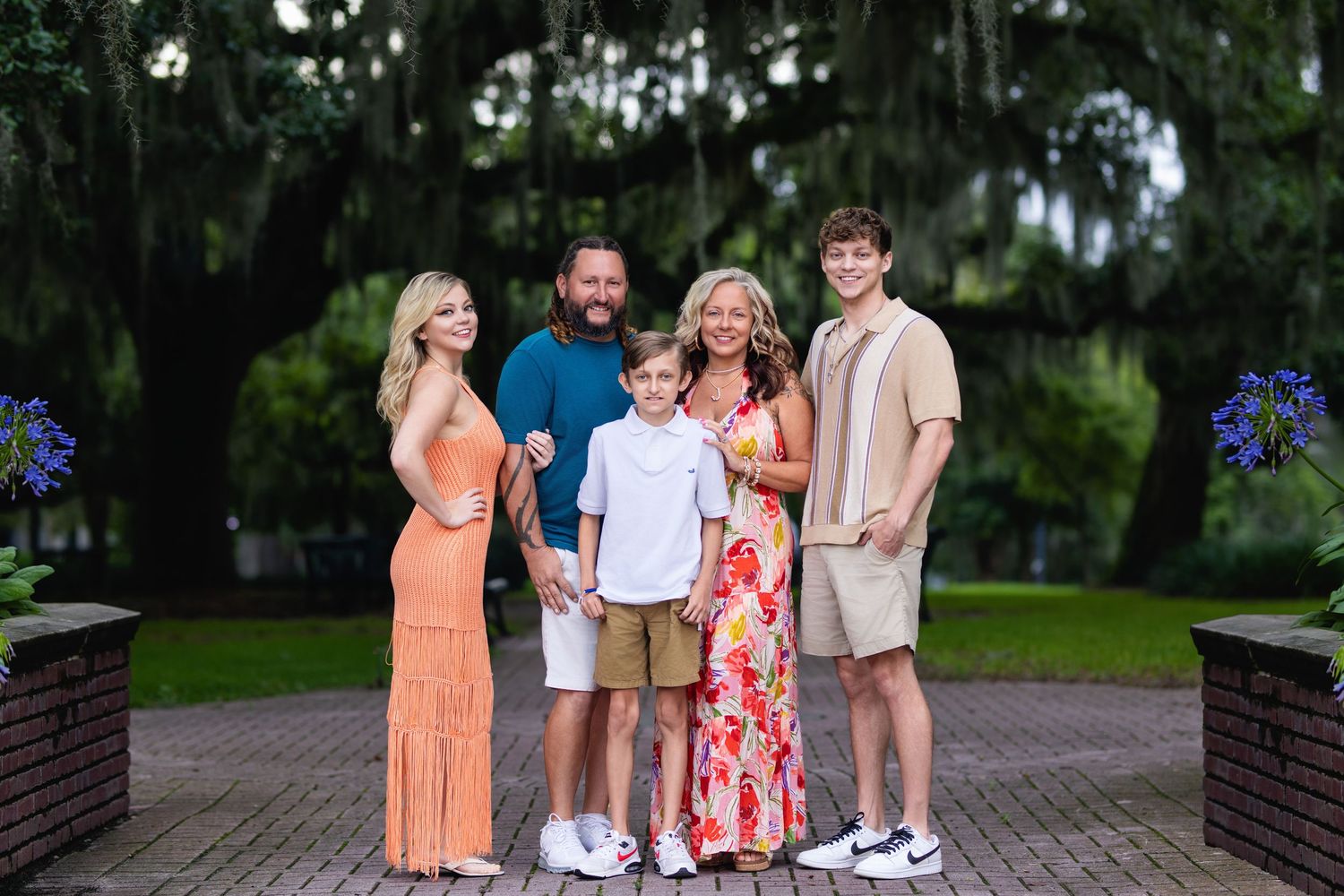 A family group poses together outdoors on a brick walkway with Spanish moss draped trees and purple flowers in the background.