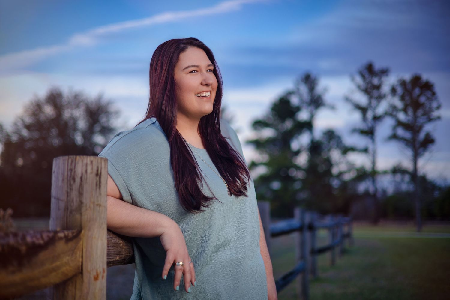 A woman in a gray shirt poses outdoors at sunset with a wooden fence and trees in the background.