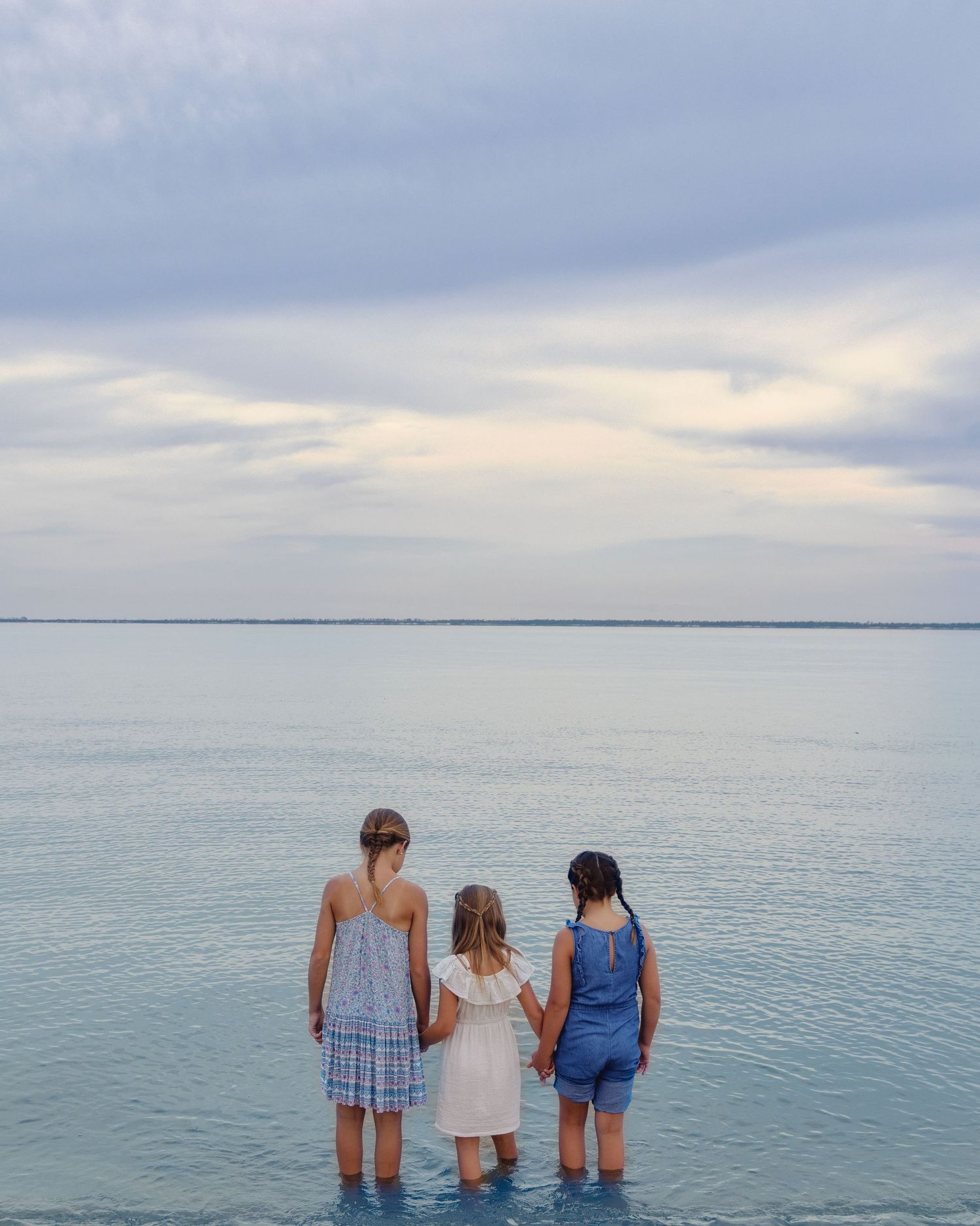 Three people in summer attire stand together looking out over a calm turquoise ocean under an overcast sky.