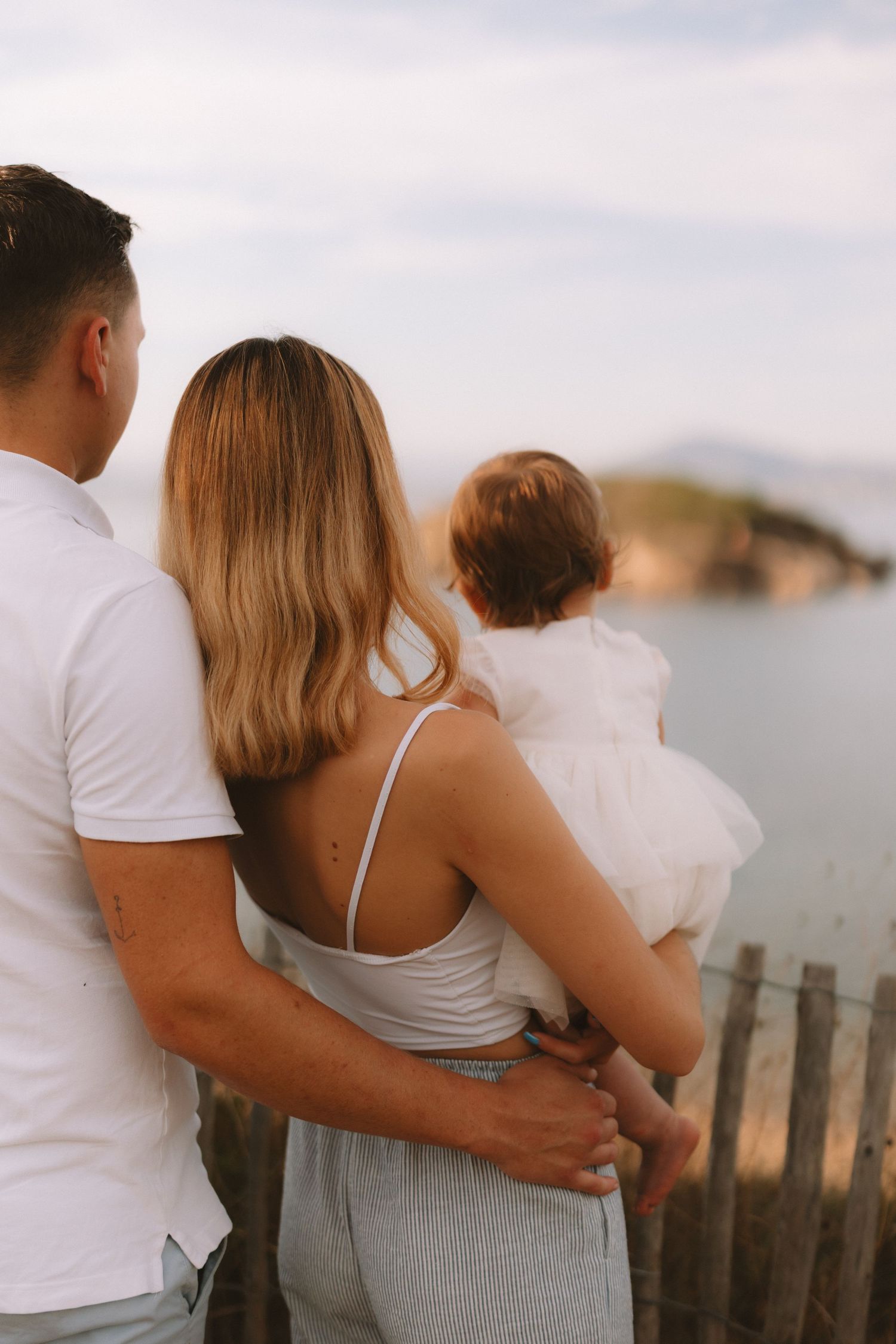 A family in white and gray clothing embraces while looking out at a scenic waterfront view during sunset.