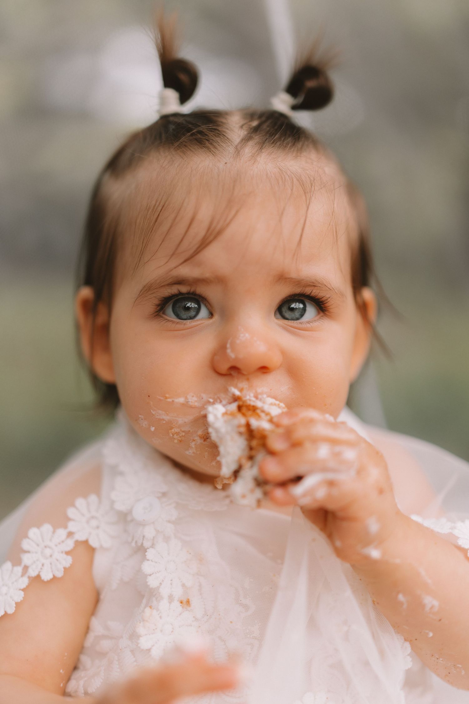 A small child in a white lacy dress enjoys birthday cake with frosting messily smeared around their mouth.