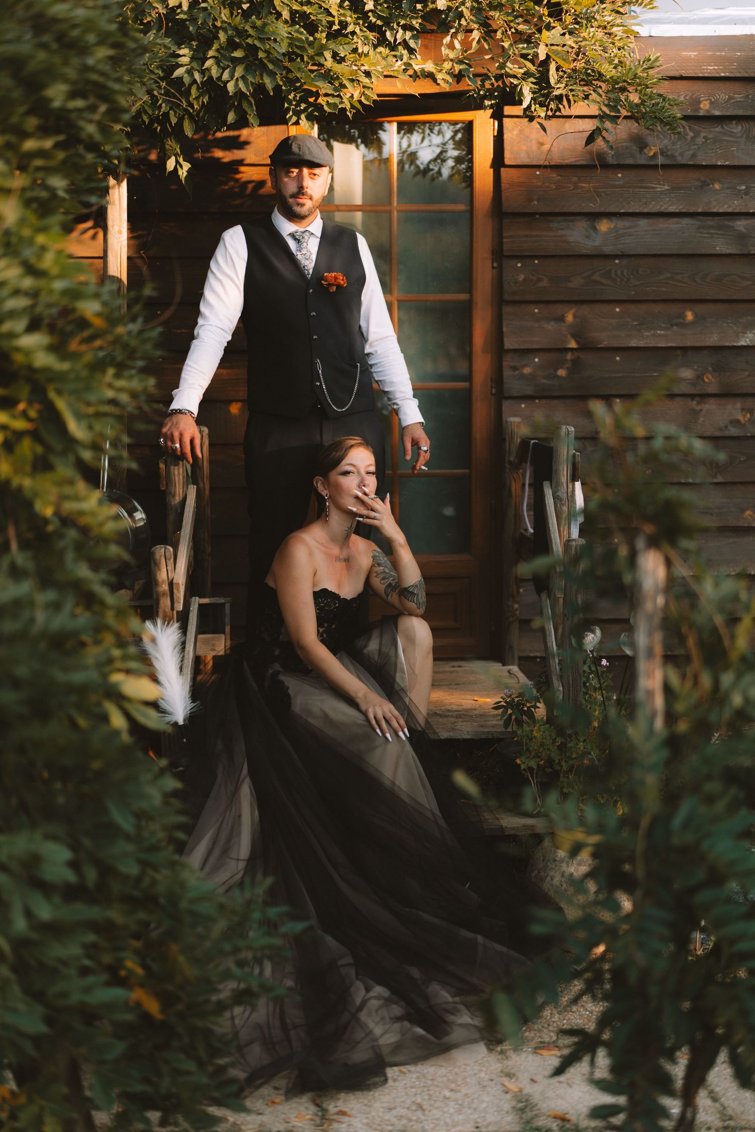 A person in Victorian-style dress sits on rustic wooden steps surrounded by green foliage in warm evening light.