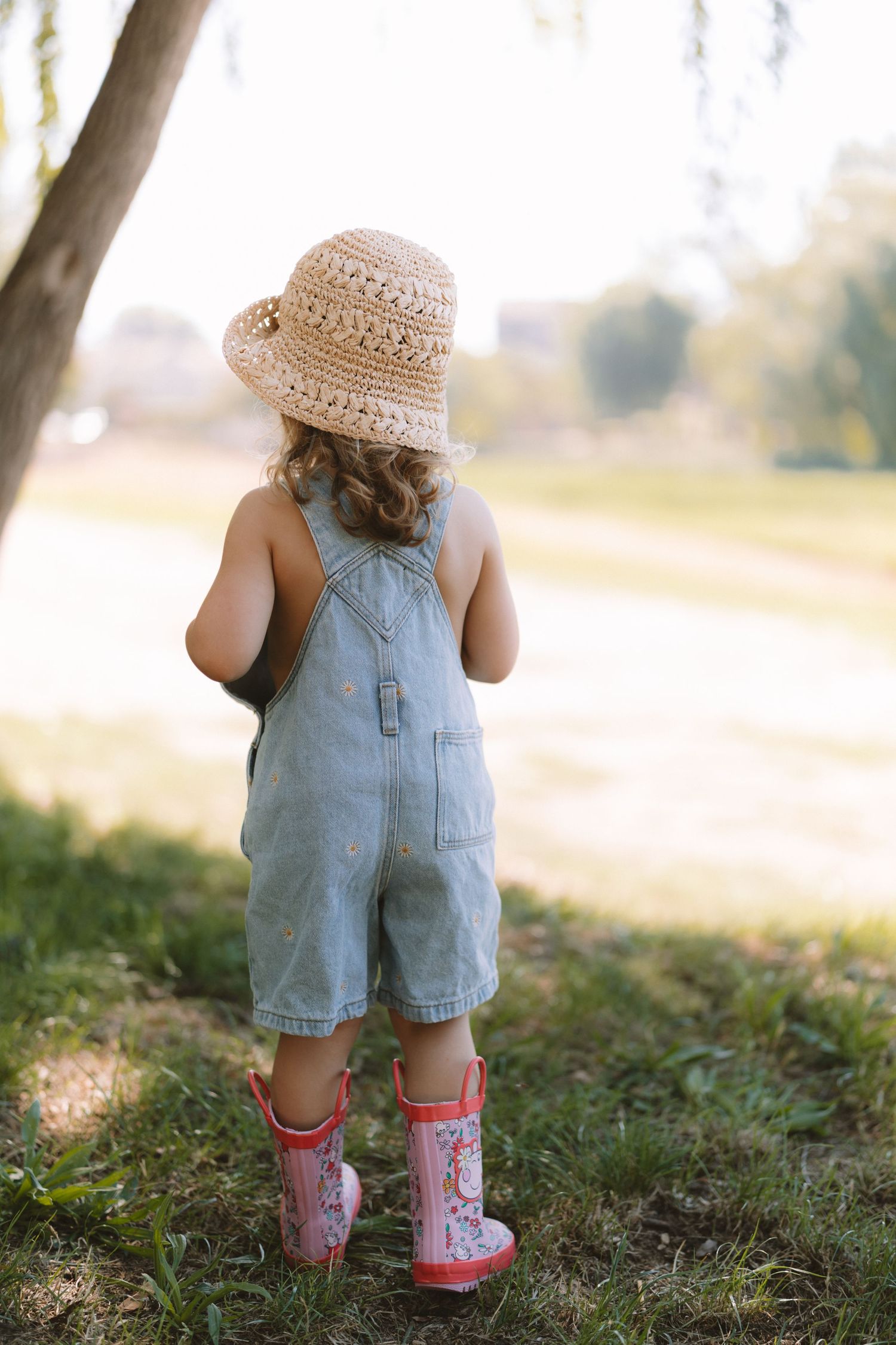 A small child wearing denim overalls and red boots stands in a grassy field on a sunny day wearing a straw hat.