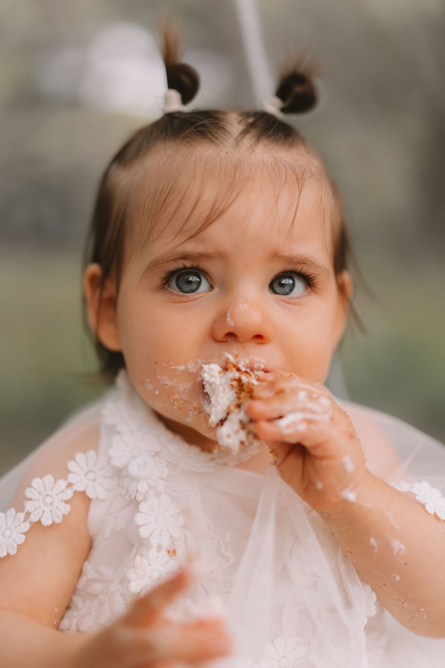 Toddler in white lace dress with pigtails enjoys cake with frosting smeared on their face.