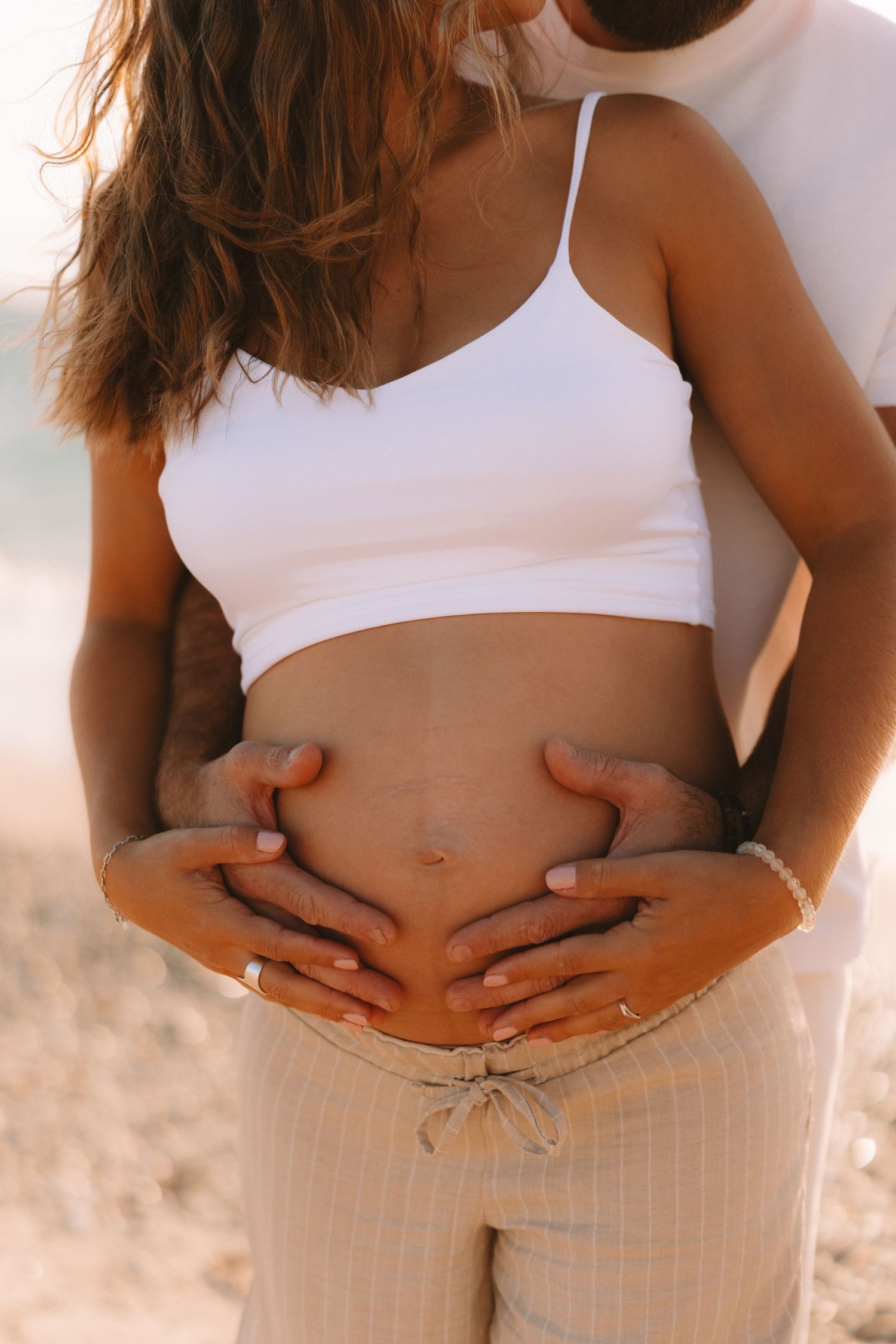 Close-up shots of a pregnant belly in white crop top and cream pants during a beach photoshoot.