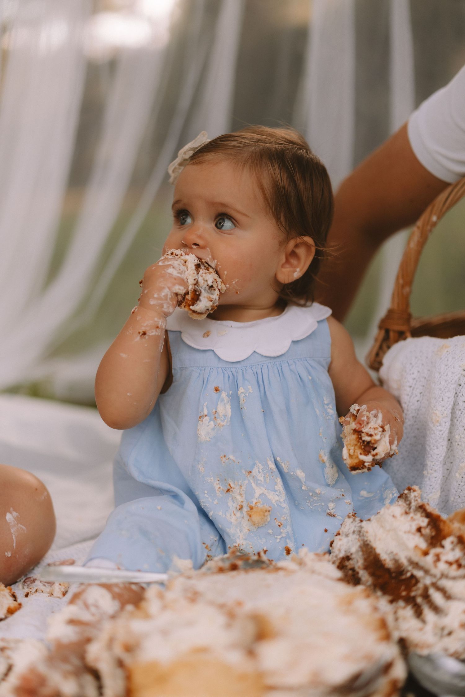 A baby in a light blue dress enjoys a messy cake smash moment with frosting all over their face and hands.