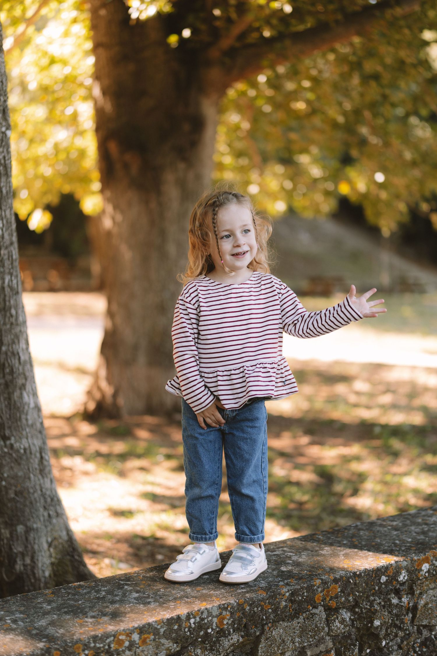 A child in a striped shirt and jeans sits on a stone wall during a golden autumn afternoon in the park.