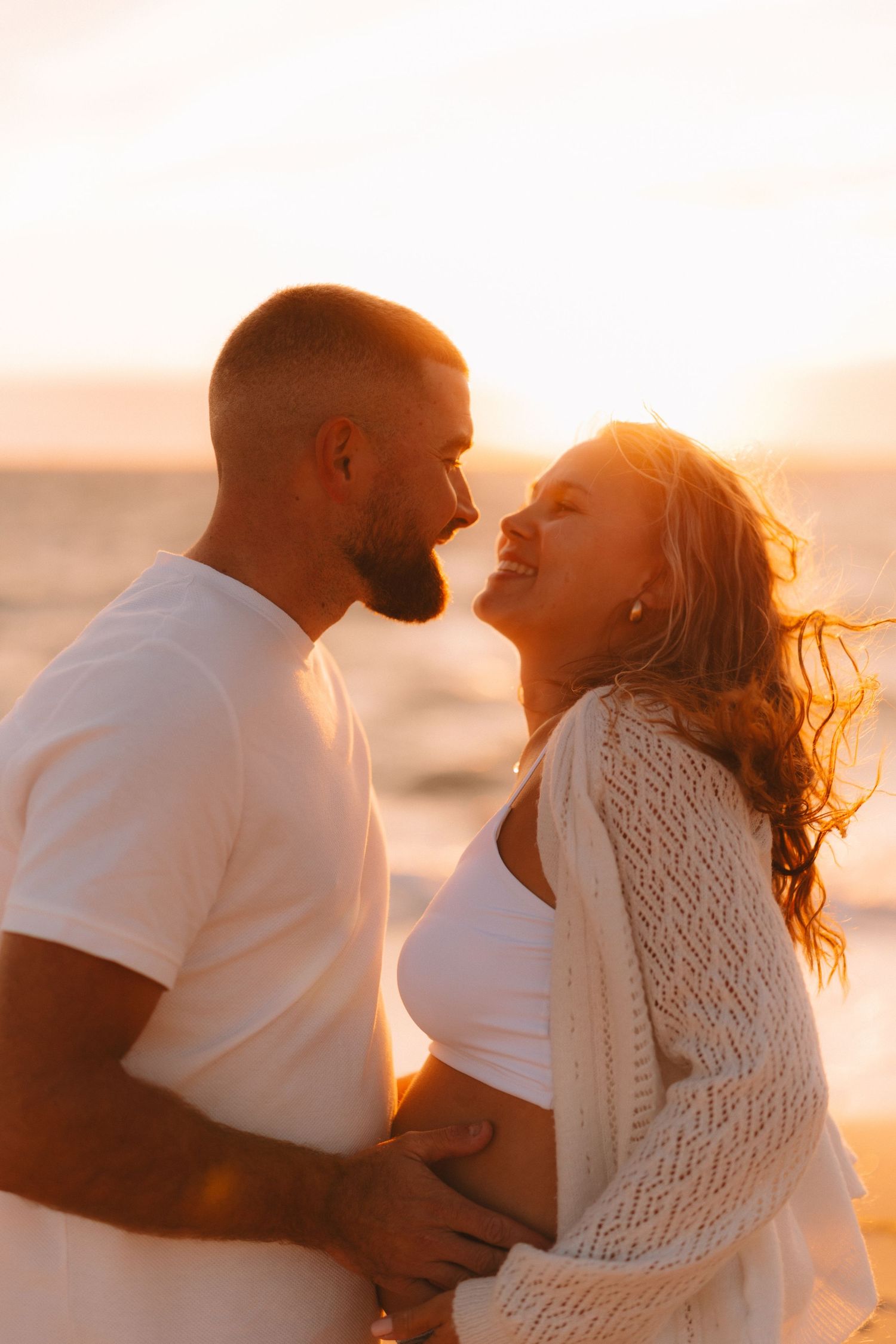 A romantic couple embraces on the beach during golden hour sunset in white outfits.