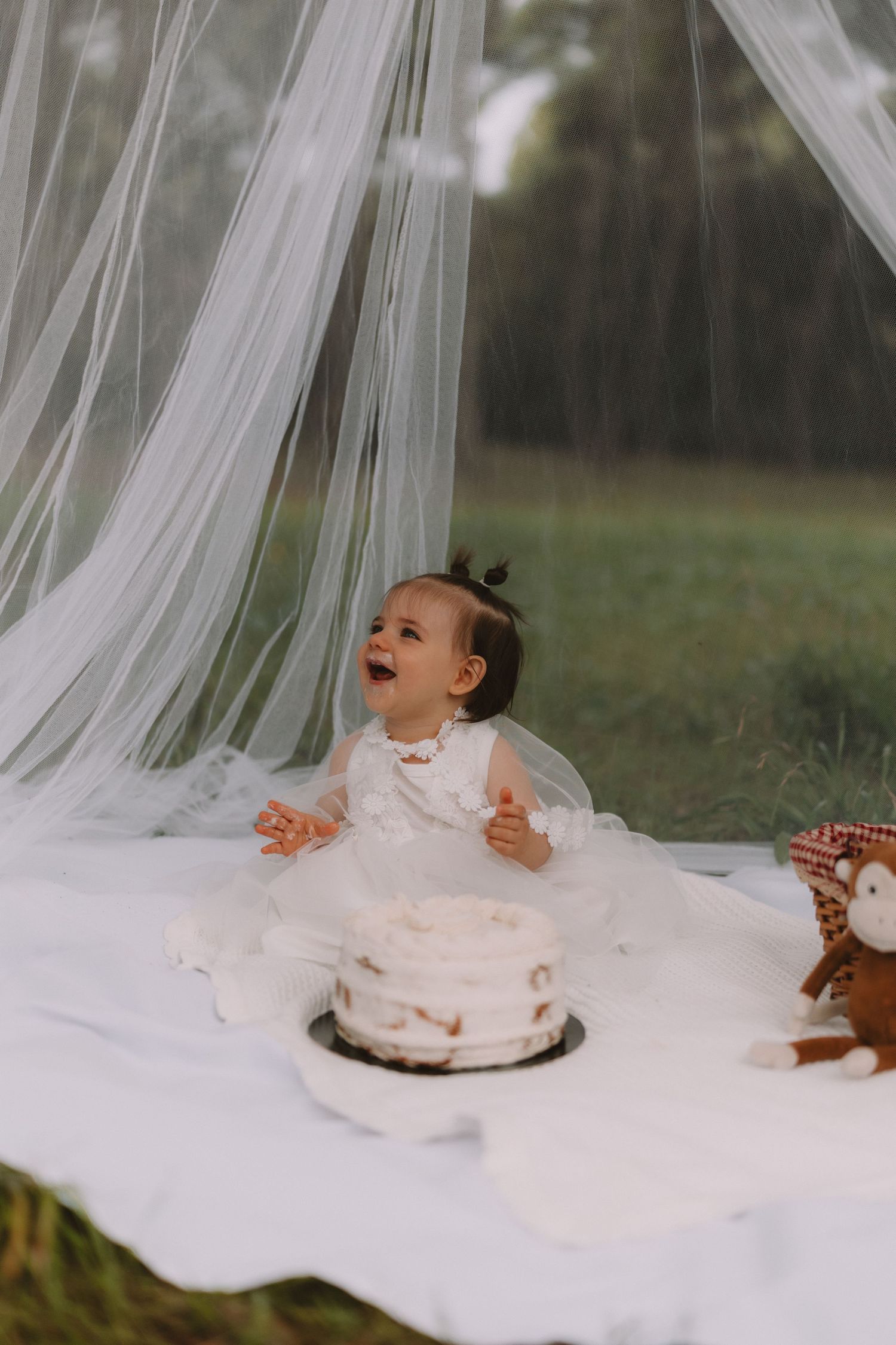 A baby in white tulle fabric laughs while playing with a smash cake during an outdoor first birthday photoshoot.