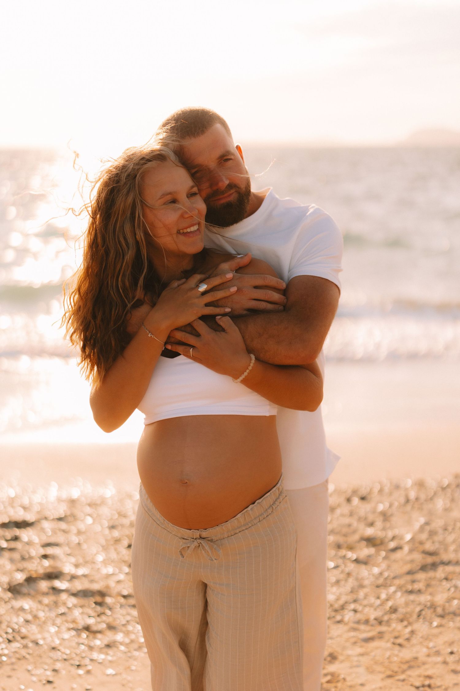 A couple shares intimate moments on a beach at sunset in matching white outfits.