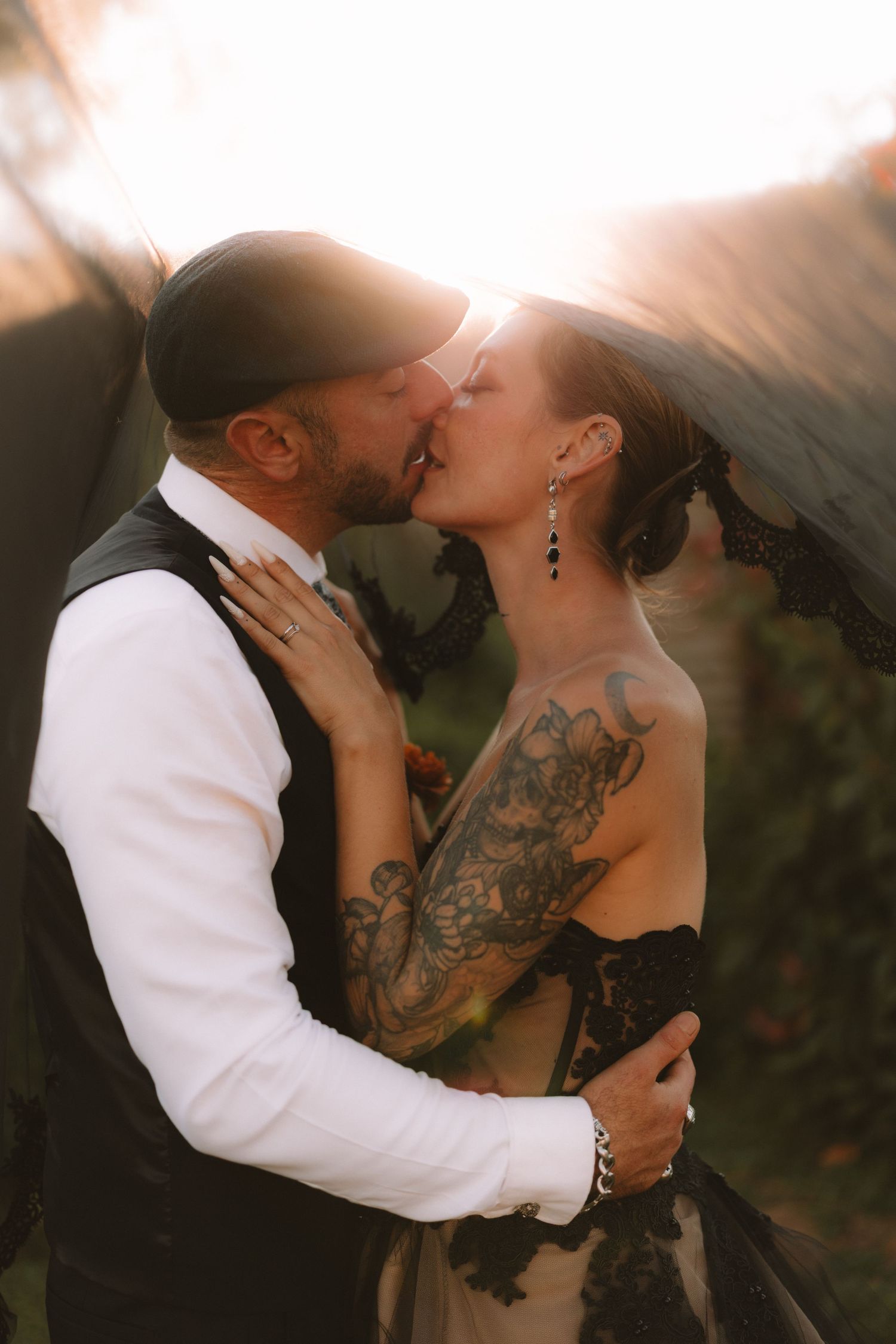 Romantic couple sharing a kiss at sunset with the bride wearing a black dress and showing elegant tattoo artwork.