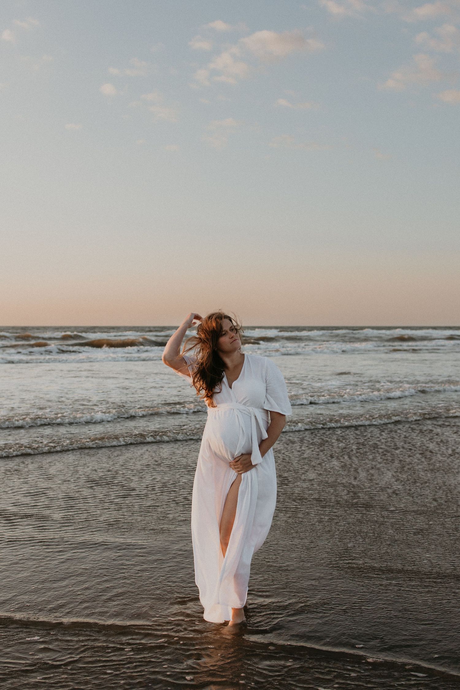 Zwangere vrouw staat rustig bij de zee tijdens golden hour, haar hand zacht op haar buik, met warm licht over het water
