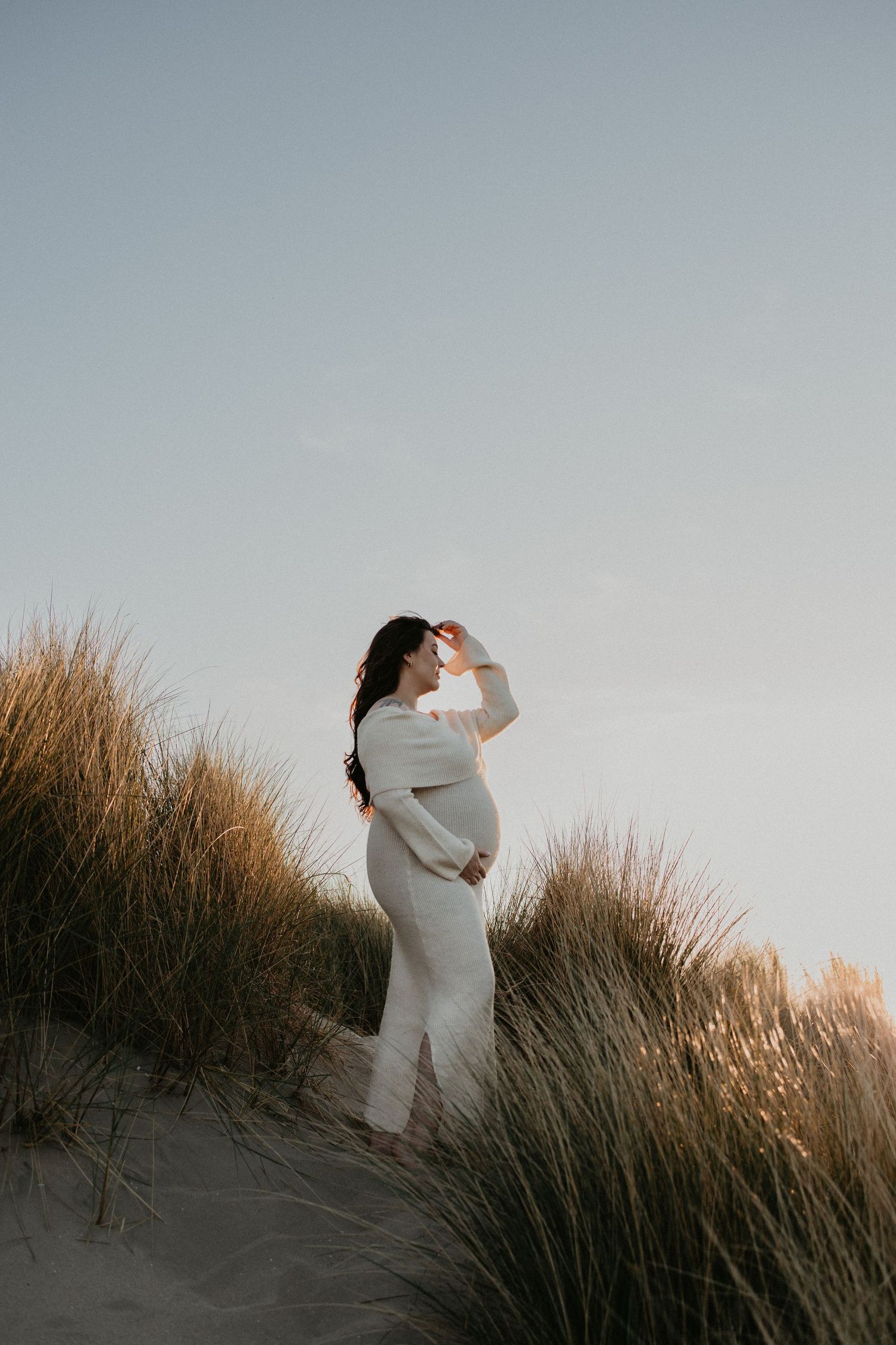 Zwangere vrouw staat alleen in de duinen tijdens golden hour, met zachte, warme zonnestralen