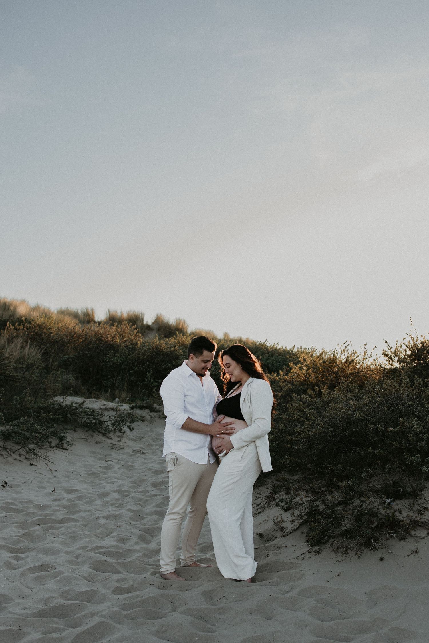 Zwanger stel staat stil in de duinen tijdens golden hour, dicht bij elkaar, in een intiem moment