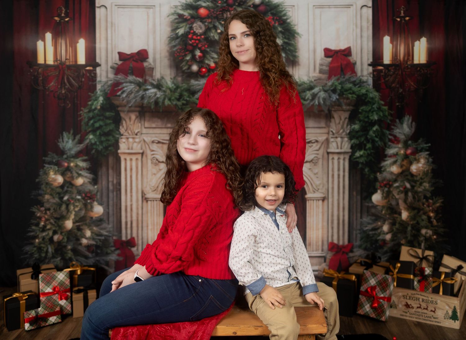A festive holiday portrait with three people wearing red sweaters by a decorated fireplace.